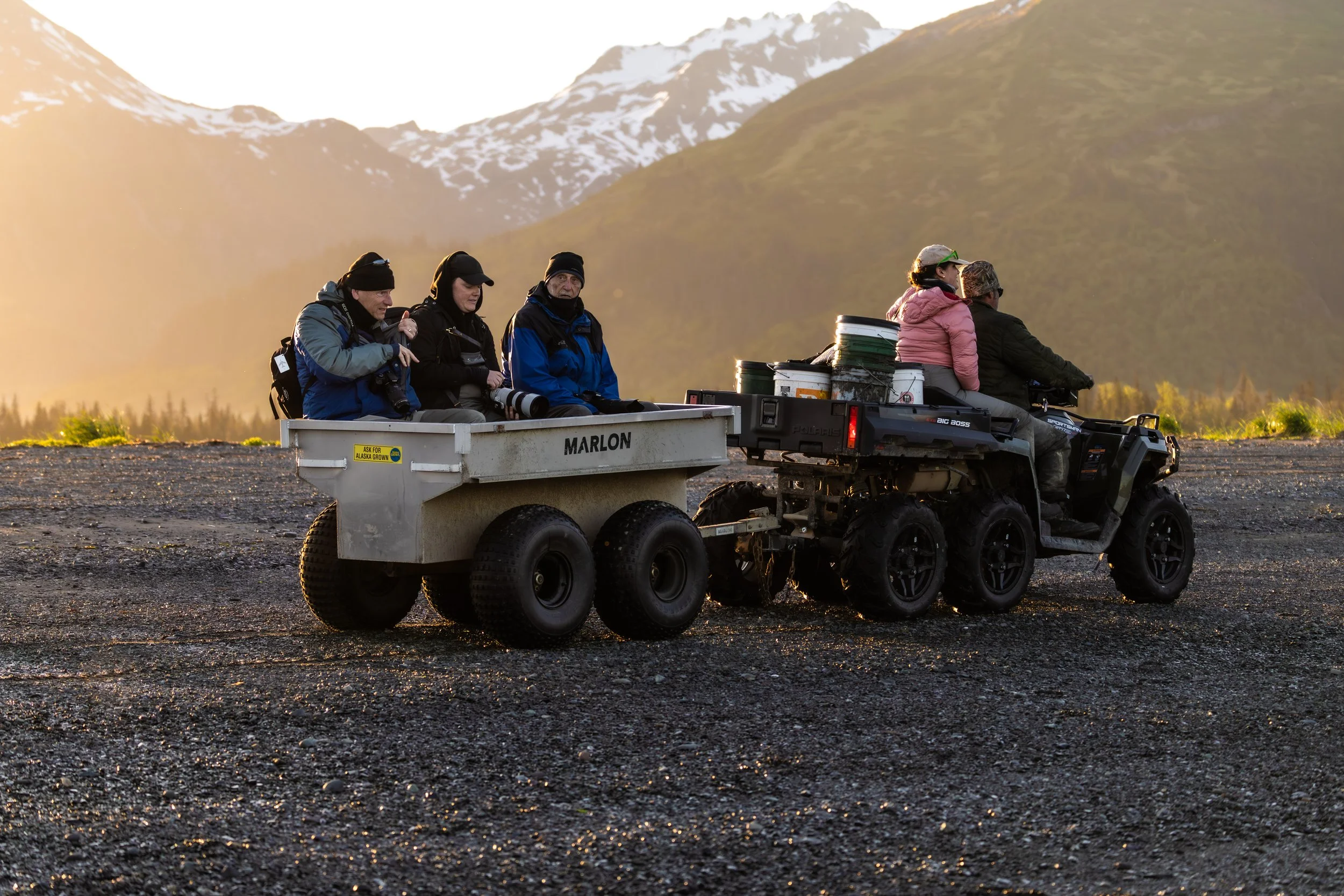 A group of people sitting on a utility trailer hitched to an ATV, with mountains in the background during sunset.