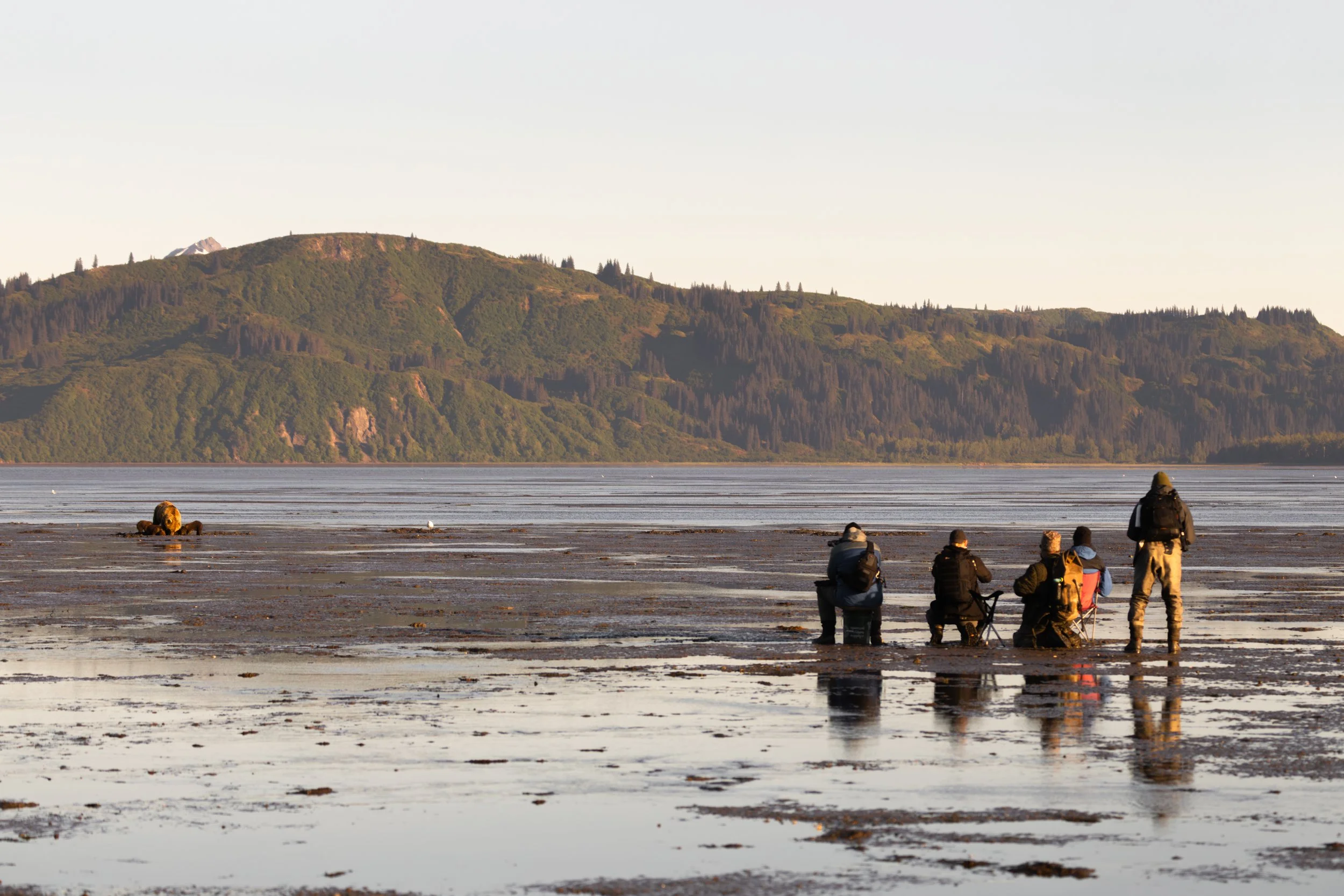 A group of people sitting and standing on a muddy beach by the water, facing a distant green mountain range under a clear sky.