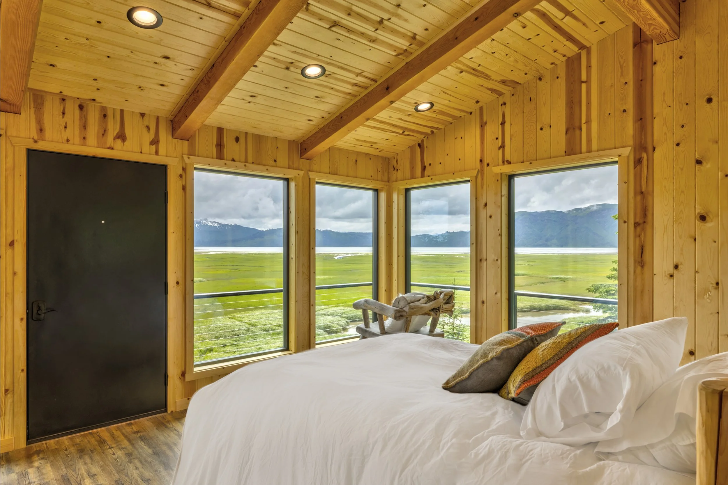 A cozy bedroom with wooden panel walls and ceiling, large windows showing a scenic view of mountains, green fields, and a cloudy sky, with a bed and pillows in the foreground.