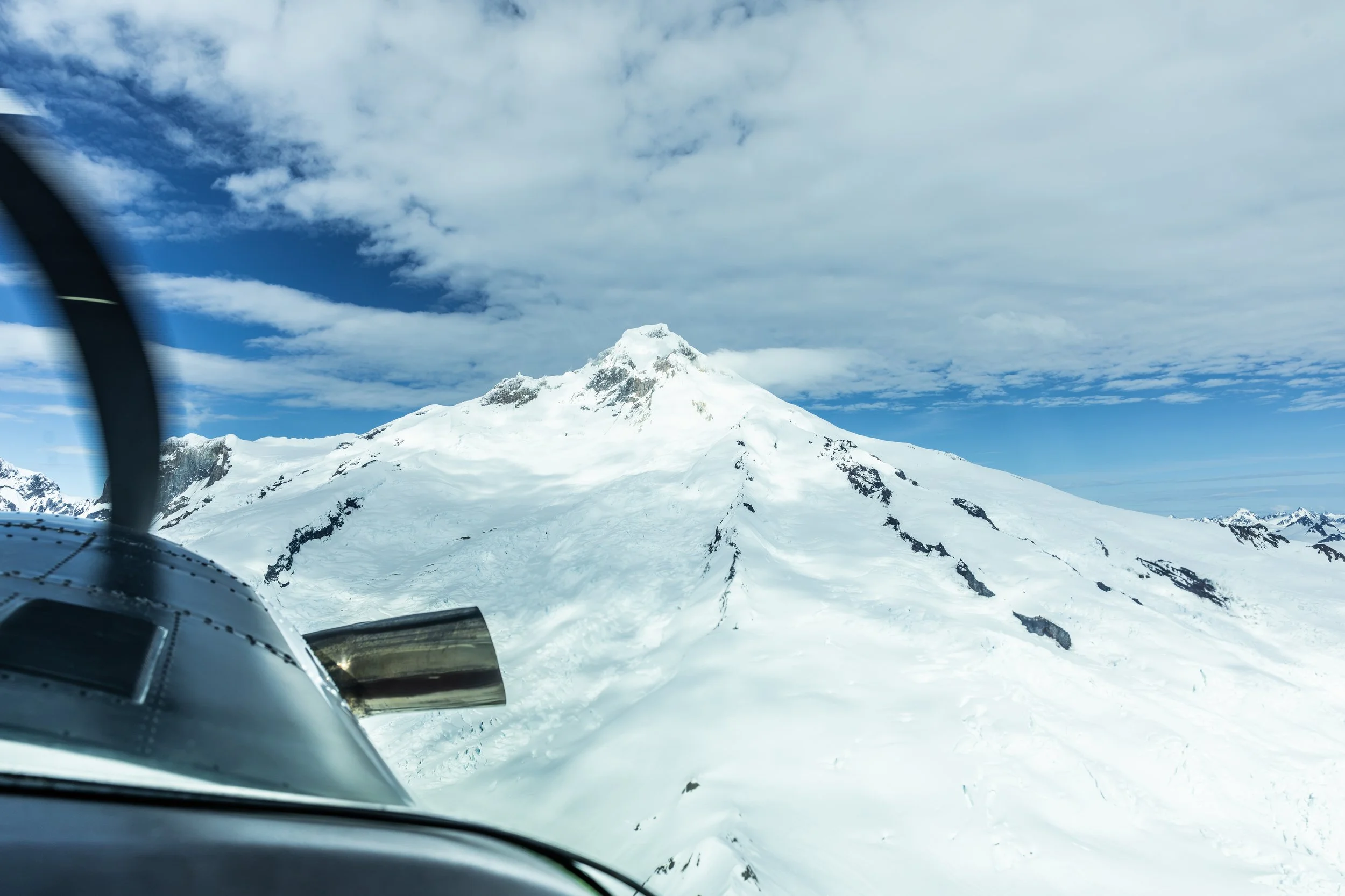 Snow-covered mountain viewed from inside an airplane or helicopter, with part of the aircraft visible in the foreground.