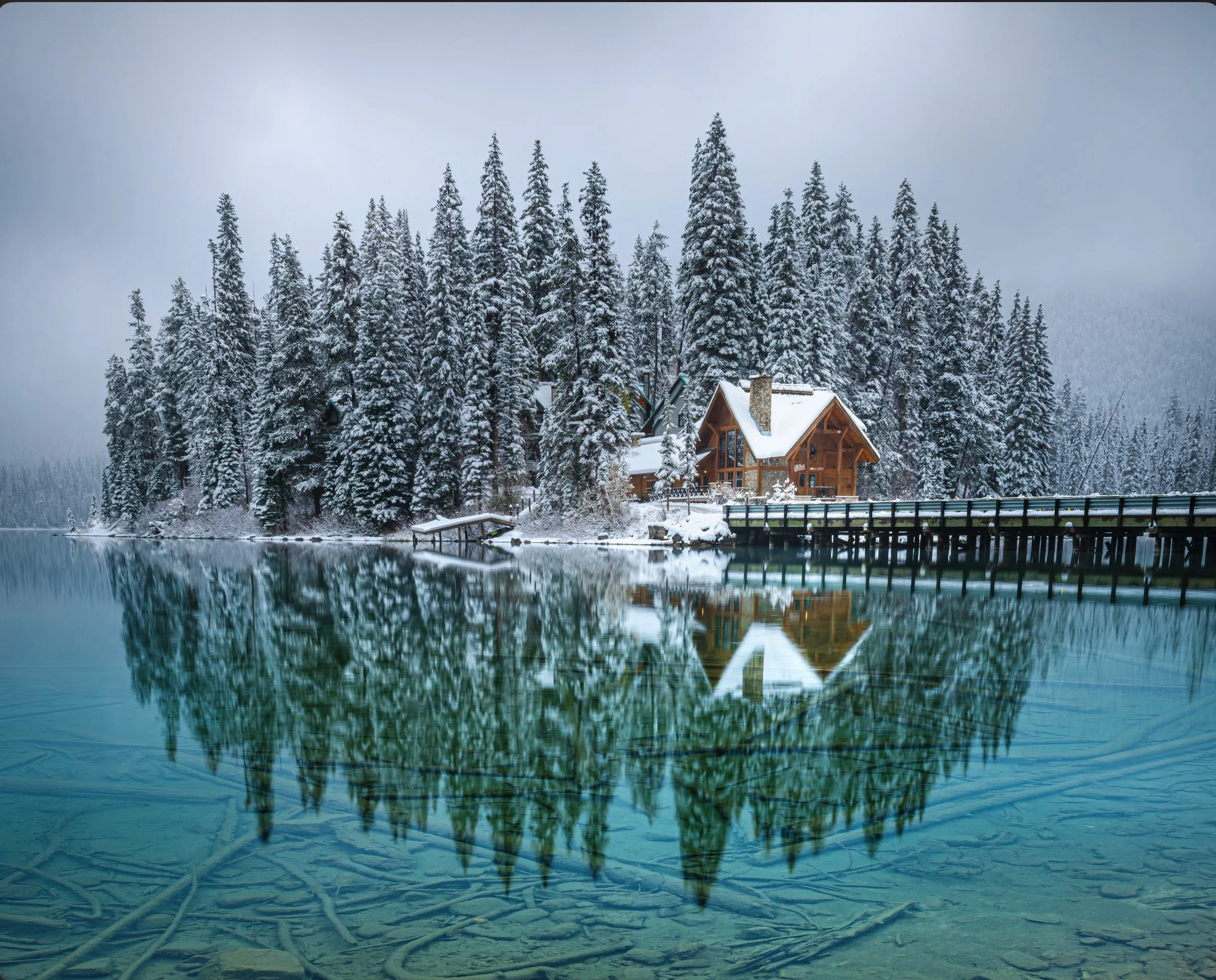 Snow-covered pine trees and a house reflected in a calm lake with a wooden dock, in a winter landscape.