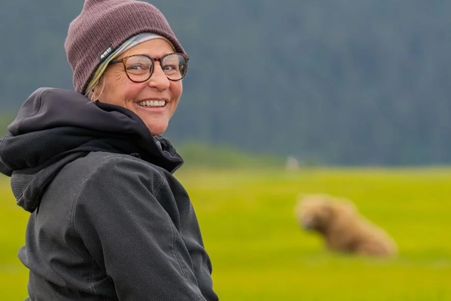A woman with glasses, wearing a maroon beanie and a black jacket, smiling outdoors with a blurred dog lying on green grass in the background.