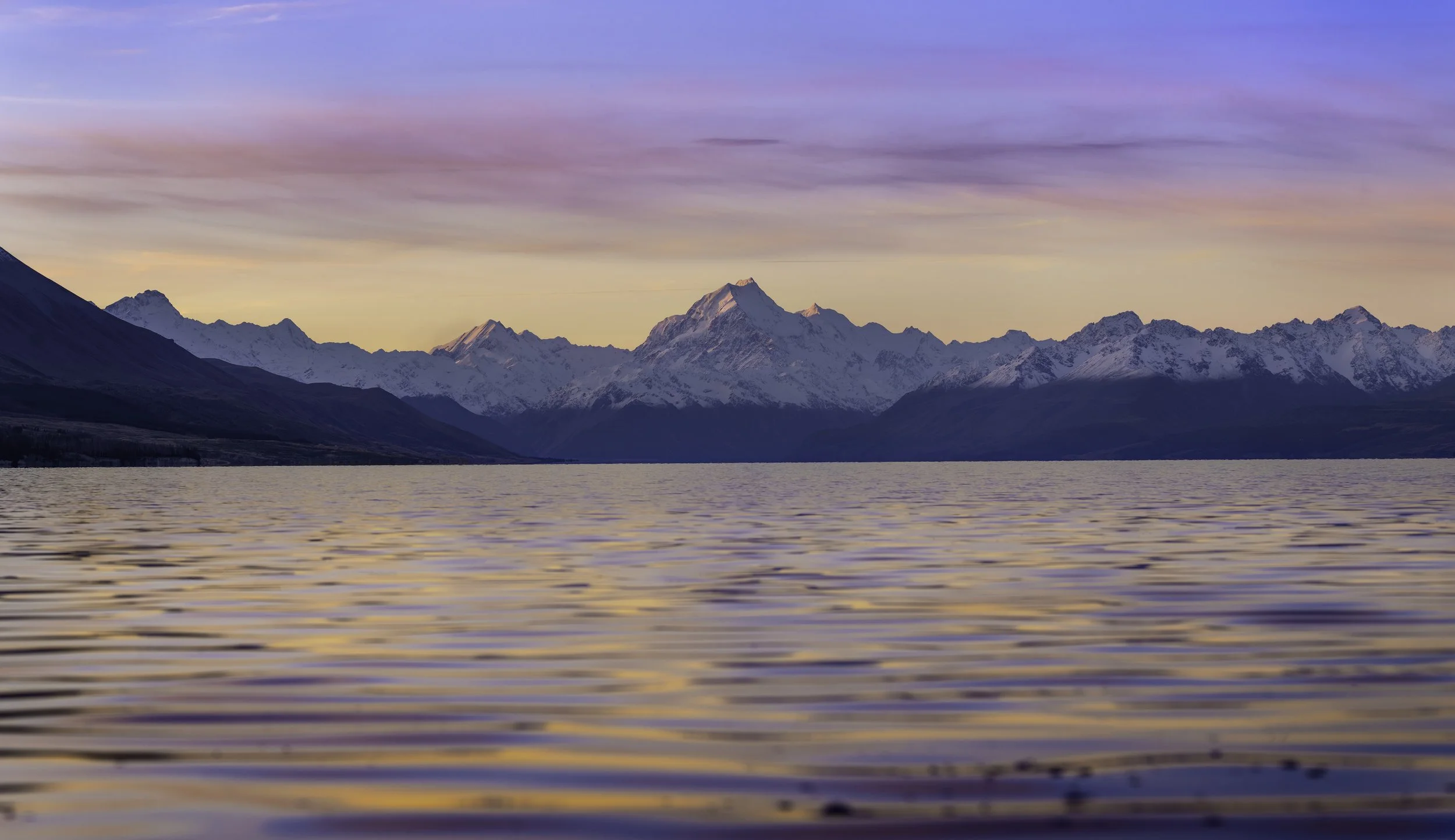 Scenic view of snow-capped mountains under a pastel-colored sky during sunset, reflected on a large body of water in the foreground.