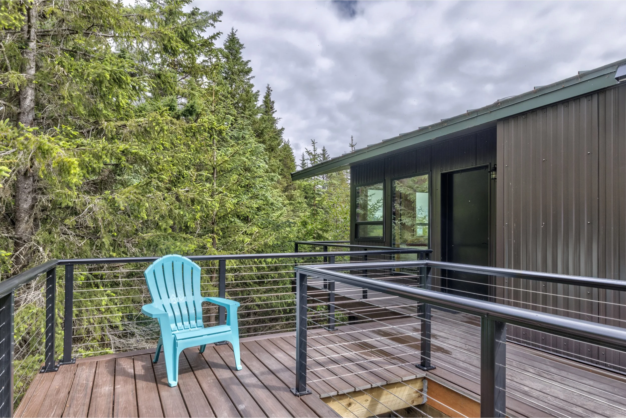 A balcony with a blue chair, wooden floor, black metal railing, adjacent to a modern house with dark exterior walls and large windows. The background features a dense forest of green trees and an overcast sky.