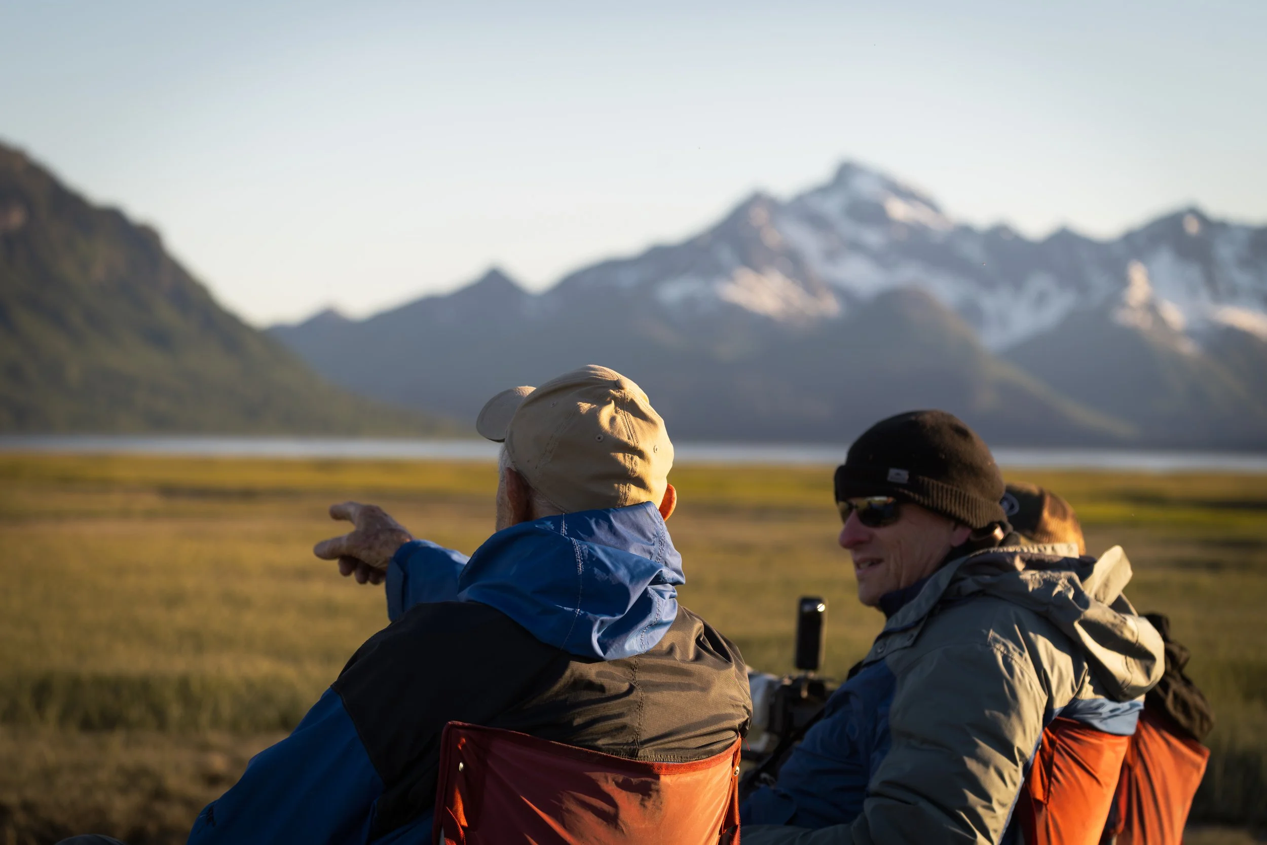 Three individuals wearing outdoor gear, sitting on a grassy plain with mountains in the background, during what appears to be late afternoon or early evening.