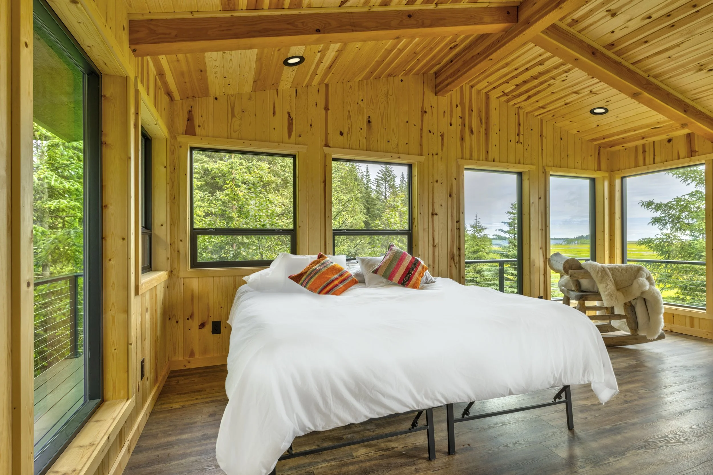 A bedroom with wooden walls and ceiling, large windows showing green trees and fields outside, a bed with white bedding and colorful pillows, and a cozy chair with a blanket.