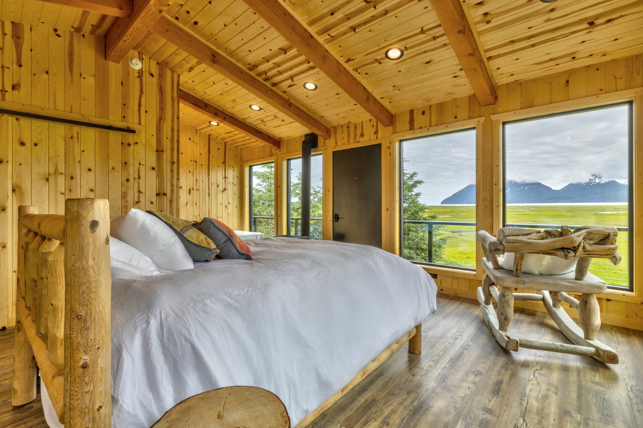 Rustic bedroom with wood-paneled walls and ceiling, large windows showing a mountain and lake landscape, a wooden bed with white bedding, and a wooden chair with a carved backrest.