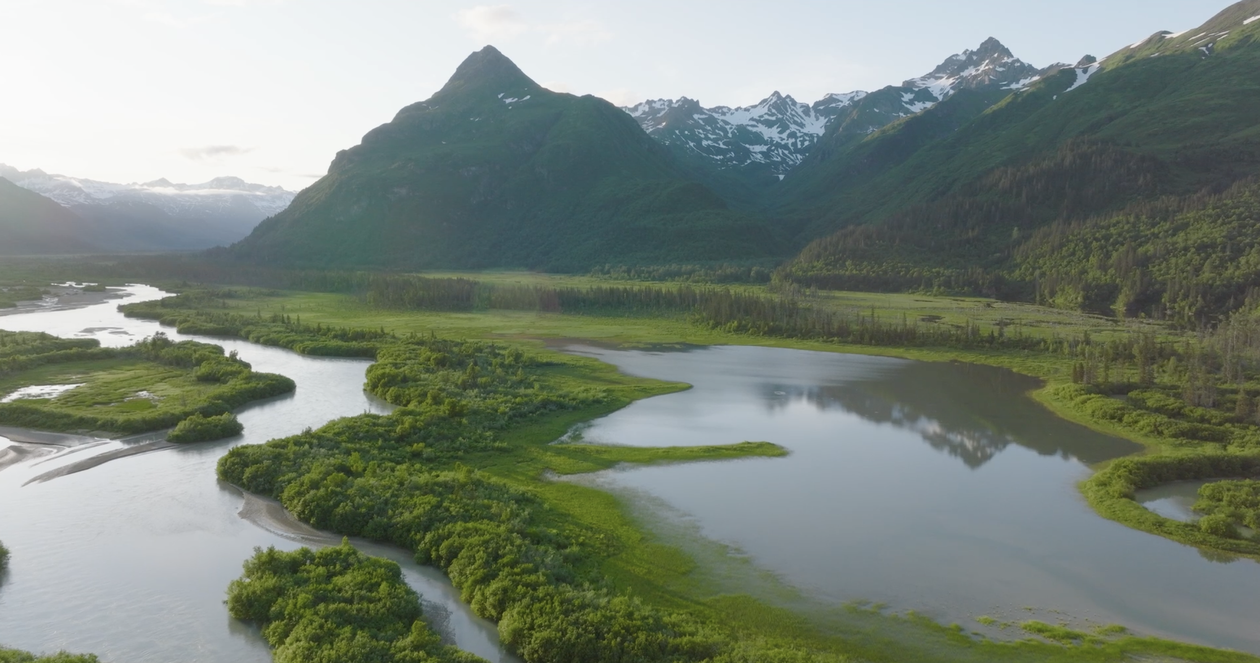 Aerial view of a winding river flowing through lush green vegetation with tall mountains in the background, some with snow.