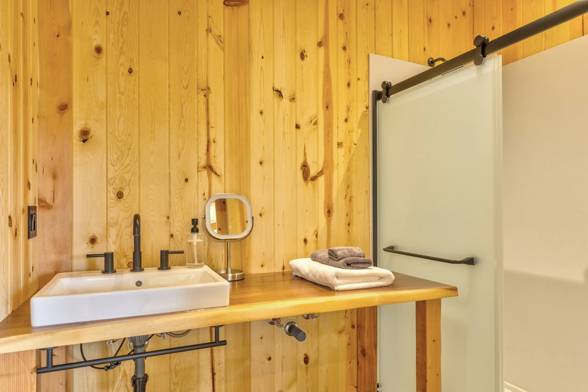 Bathroom with wood-paneled wall, white sink, black faucet, small mirror, soap dispenser, white folded towels, and a sliding shower door with black hardware.