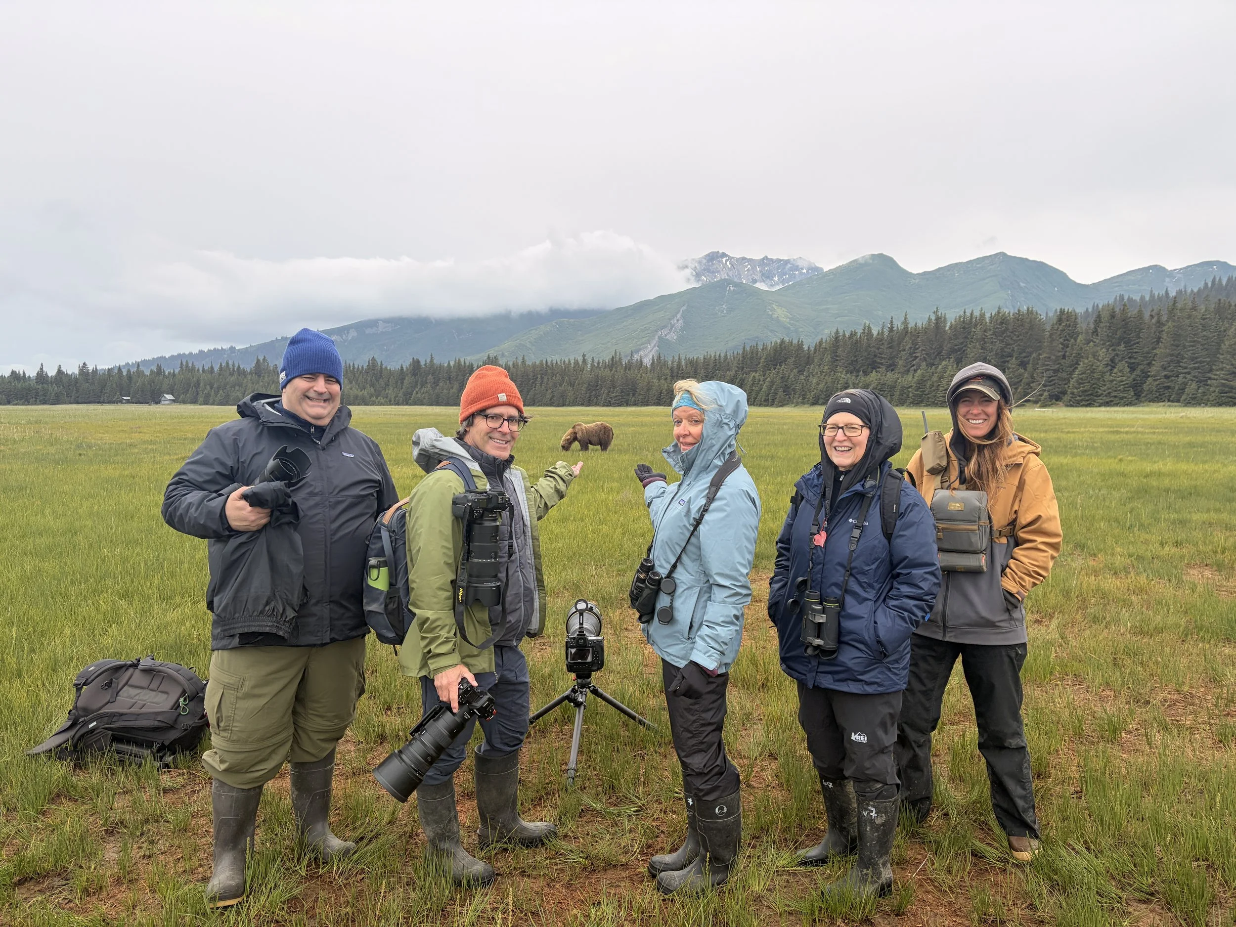 Group of five people standing in a grassy field with mountains in the background. They are dressed in outdoor gear, some holding cameras, and are smiling. There is a bison in the distance and cloudy skies.