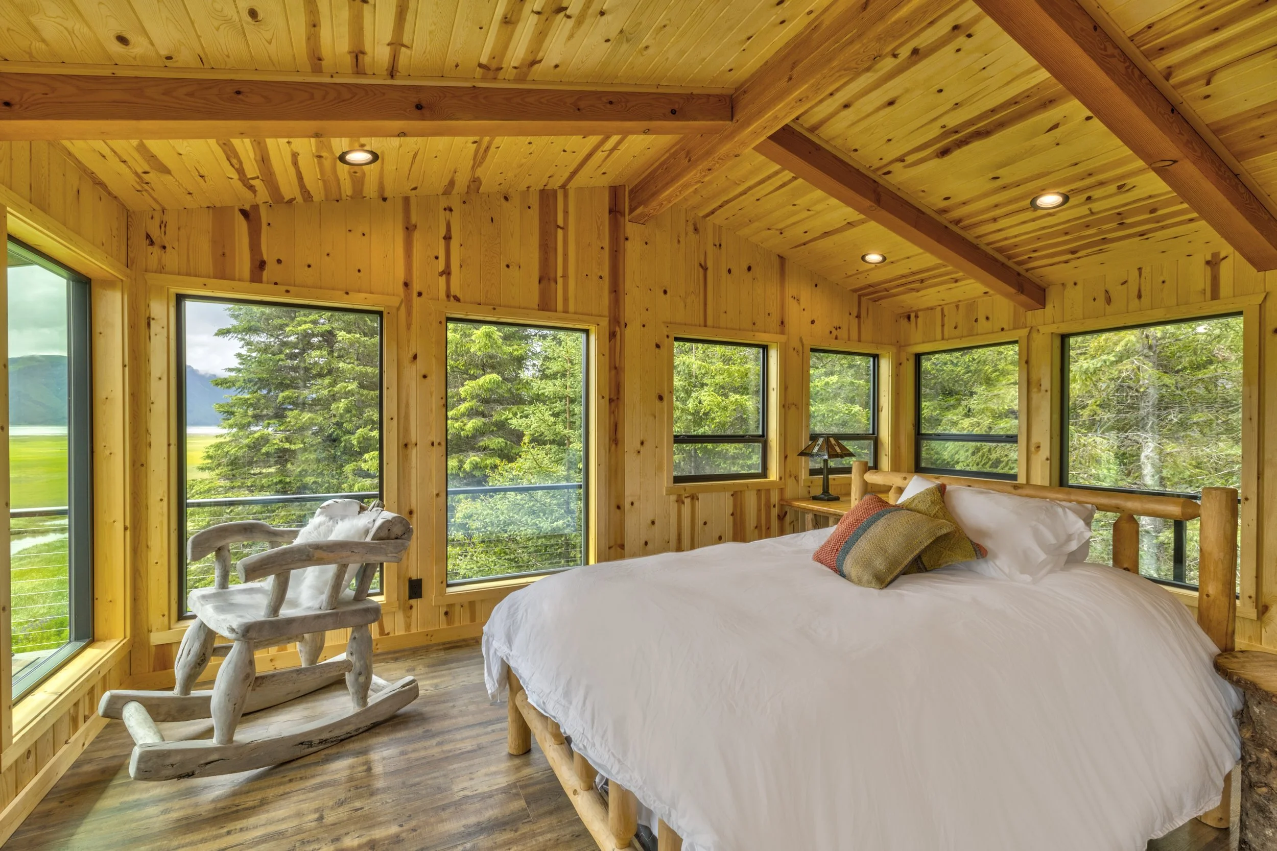 A rustic bedroom with wooden walls and ceiling, large windows showing green trees outside, a bed with white bedding and colorful pillows, a wooden rocking chair with a plush animal on it, and a small table with a lamp.