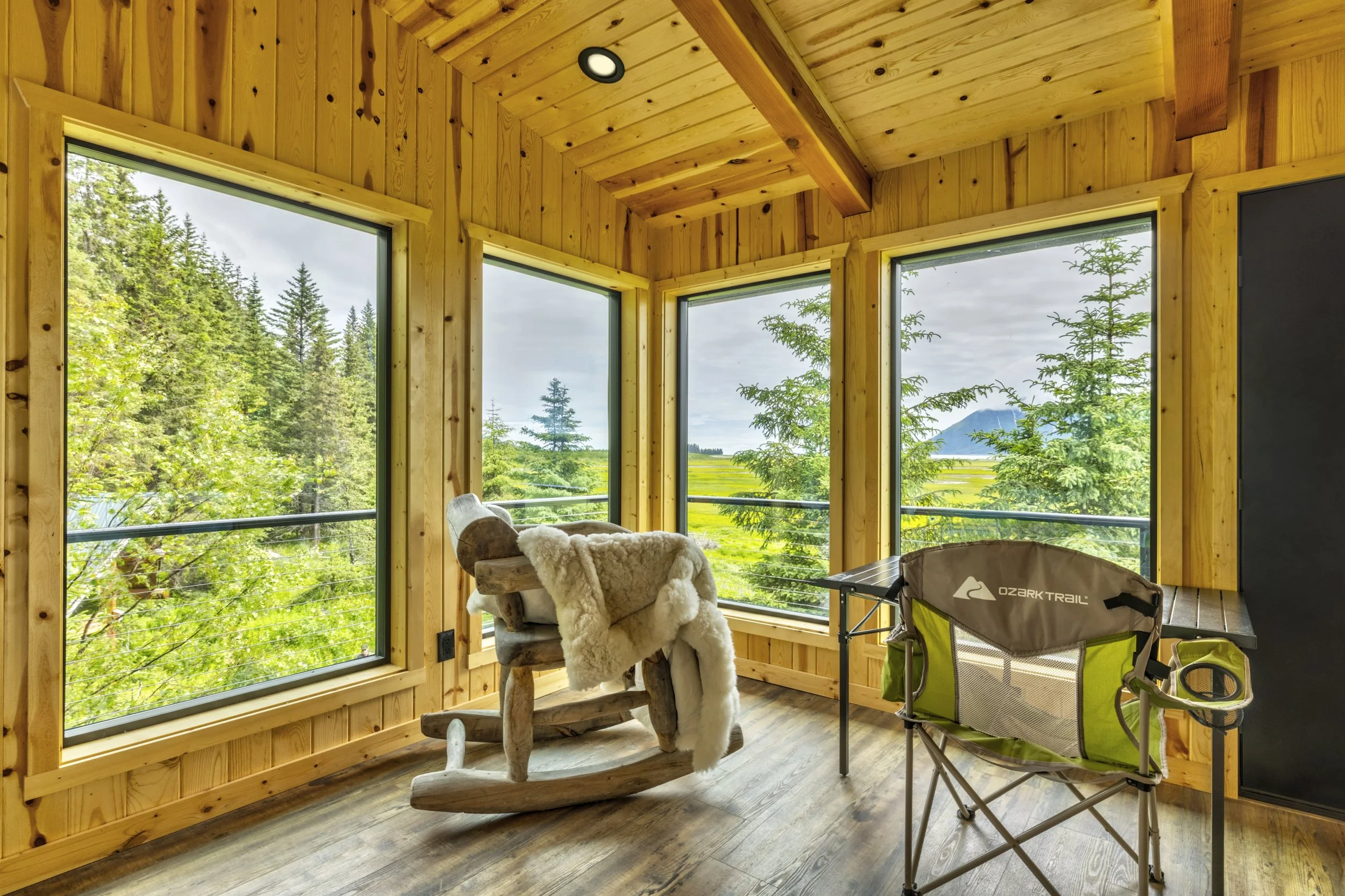 A cozy corner in a wooden cabin with large windows overlooking a lush green forest and distant mountains. The space features a rustic wooden rocking chair draped with a sheepskin, a green camping chair, and a small table, creating a comfortable outdo