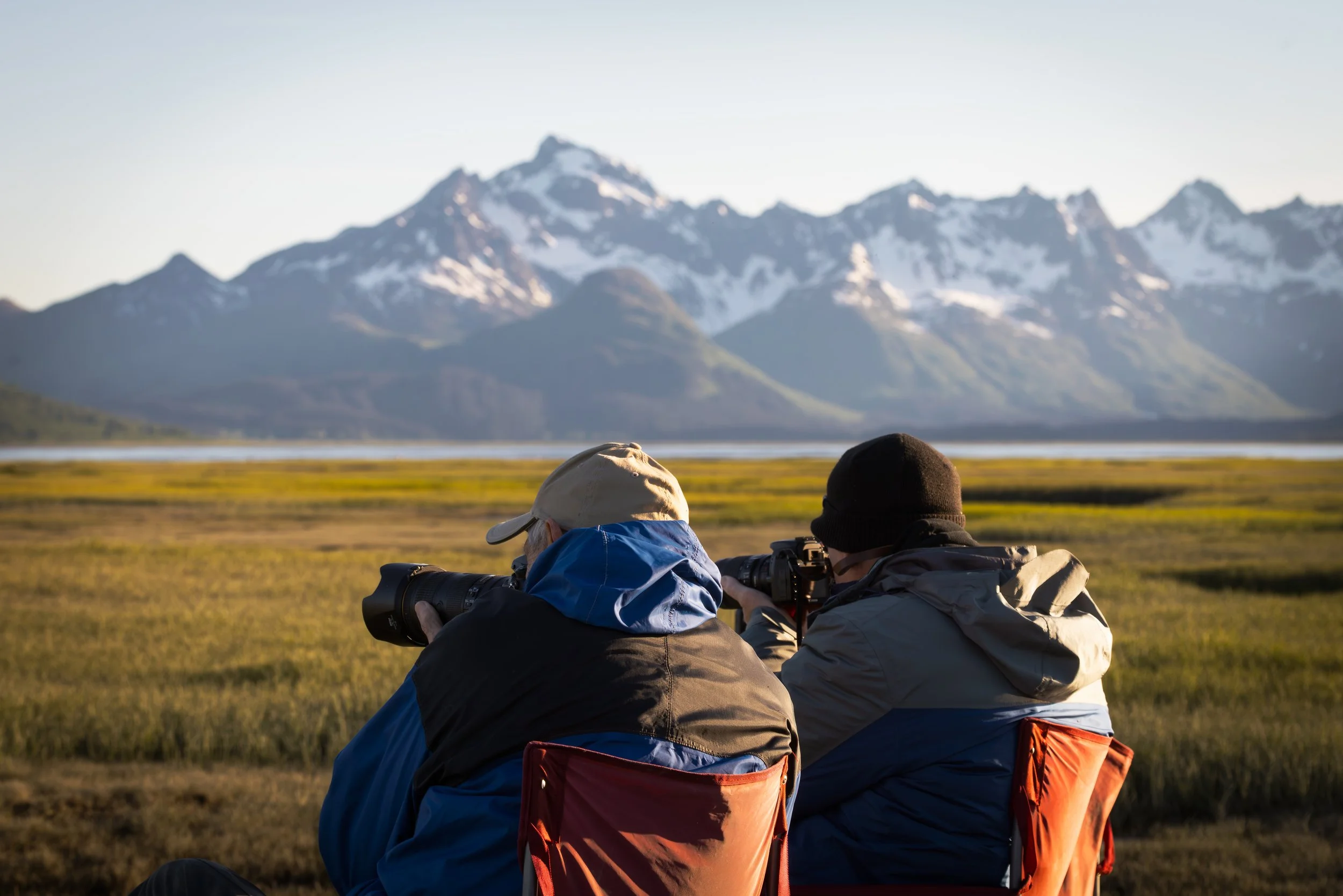 Two photographers sitting outdoors in front of a mountain range, taking photos of snow-capped peaks during daylight, with a grassy plain in the foreground.