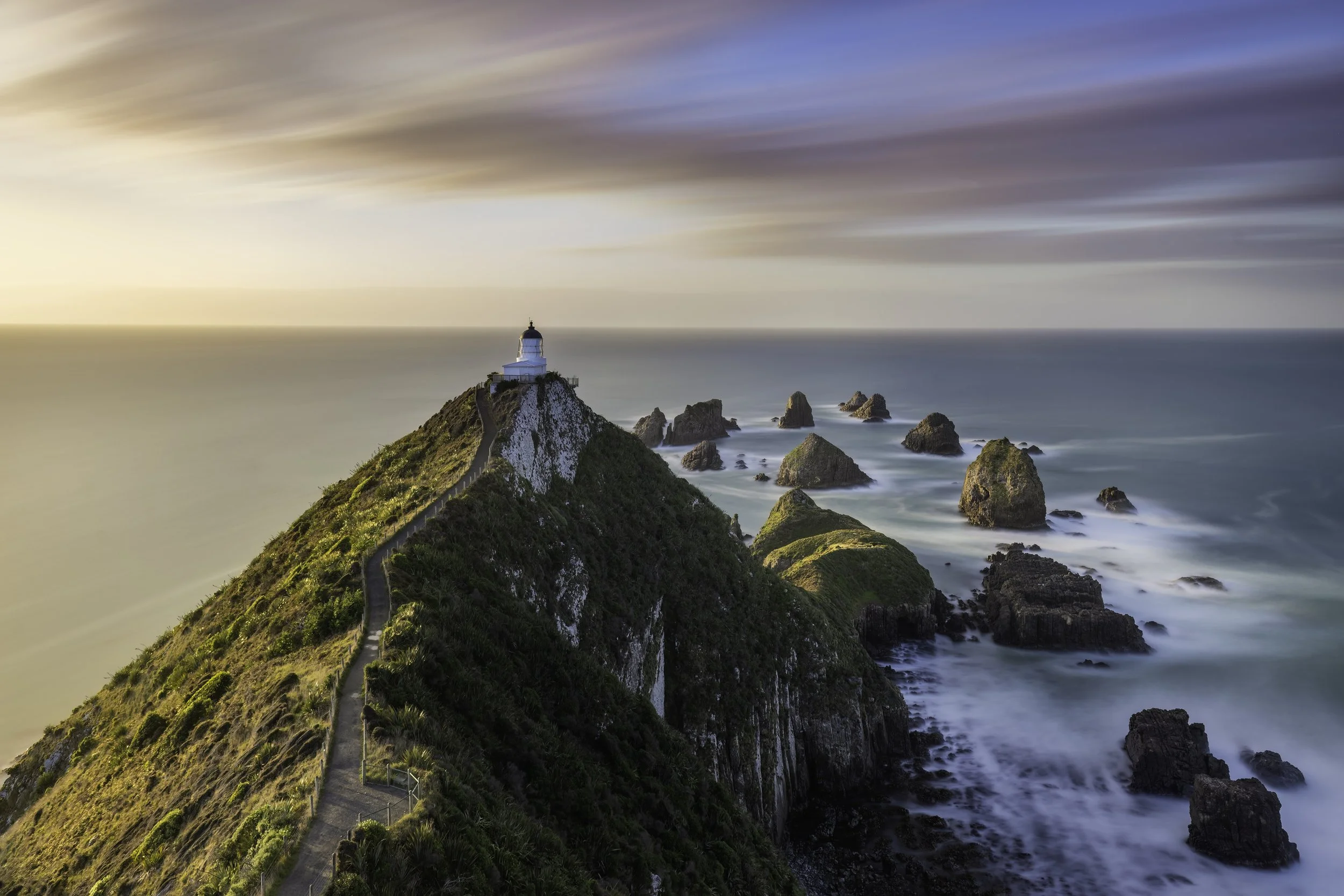 A lighthouse on a cliff overlooking the ocean during sunset, with clouds streaking across the sky.