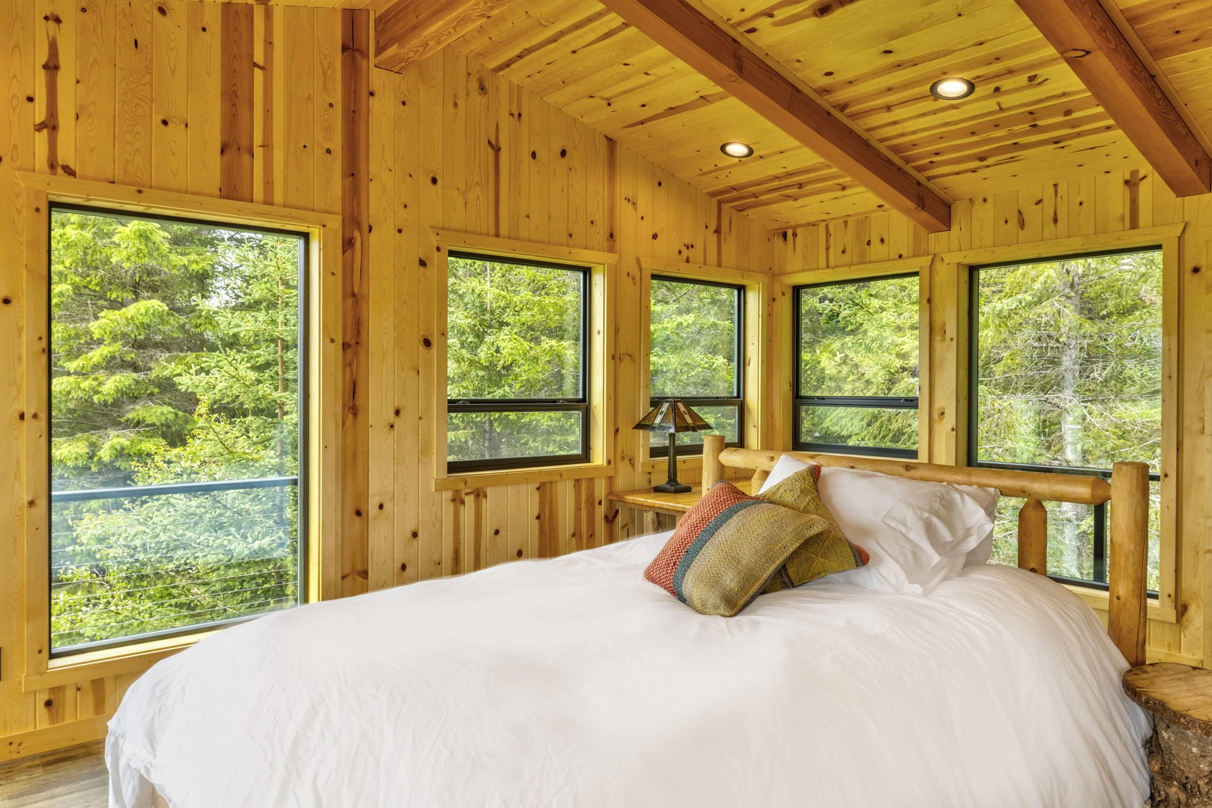 A cozy bedroom with wooden walls, ceiling, and furniture. There are four large windows showing green trees outside. The bed has white bedding and colorful pillows, and a small bedside lamp is visible.