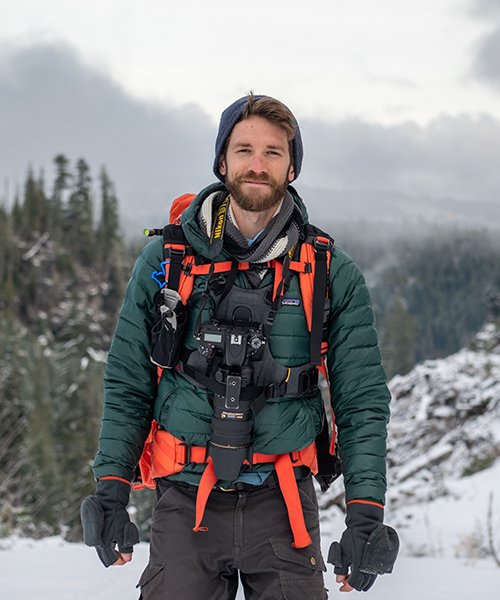 Man hiking in snowy, mountainous terrain wearing a blue beanie, green jacket, and carrying a camera with a strap, with trees and snow-covered landscape in the background.