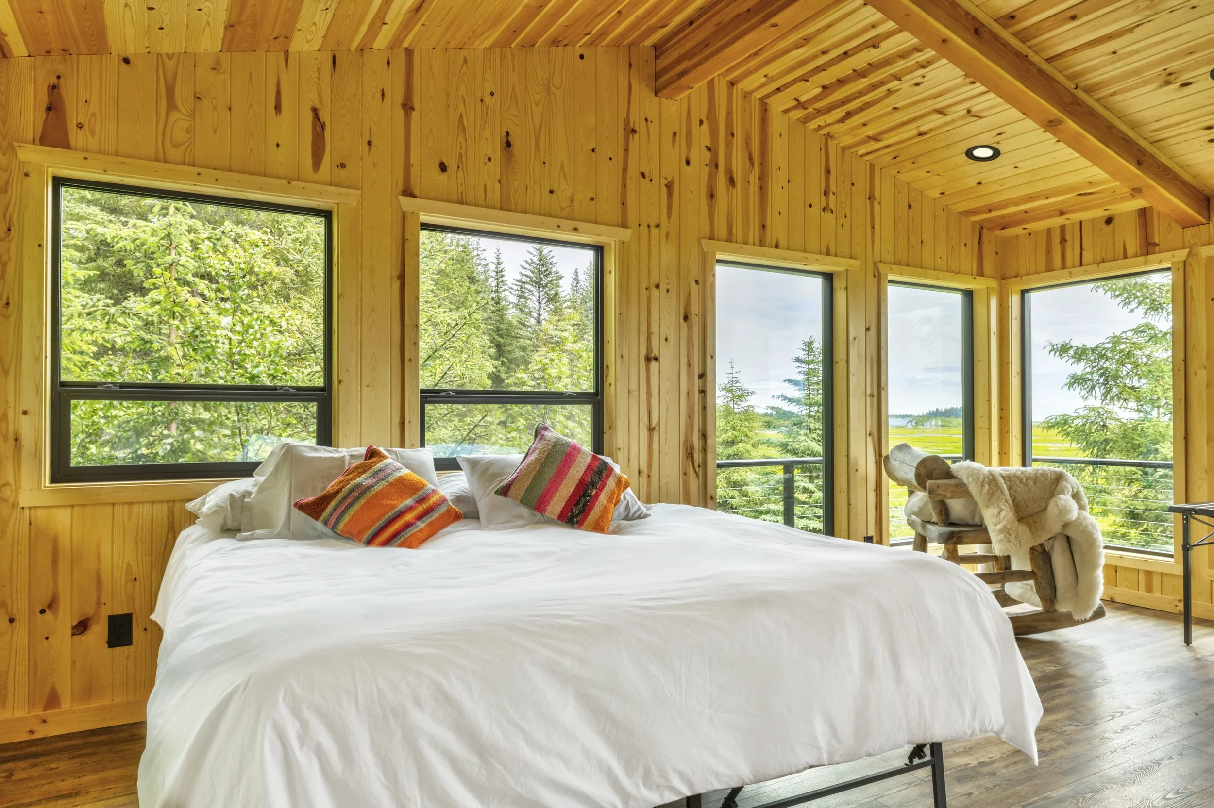A cozy bedroom with wooden walls and ceiling, large windows showing greenery and trees outside, a white bed with colorful striped pillows, and a rustic wooden chair with a sheepskin throw.