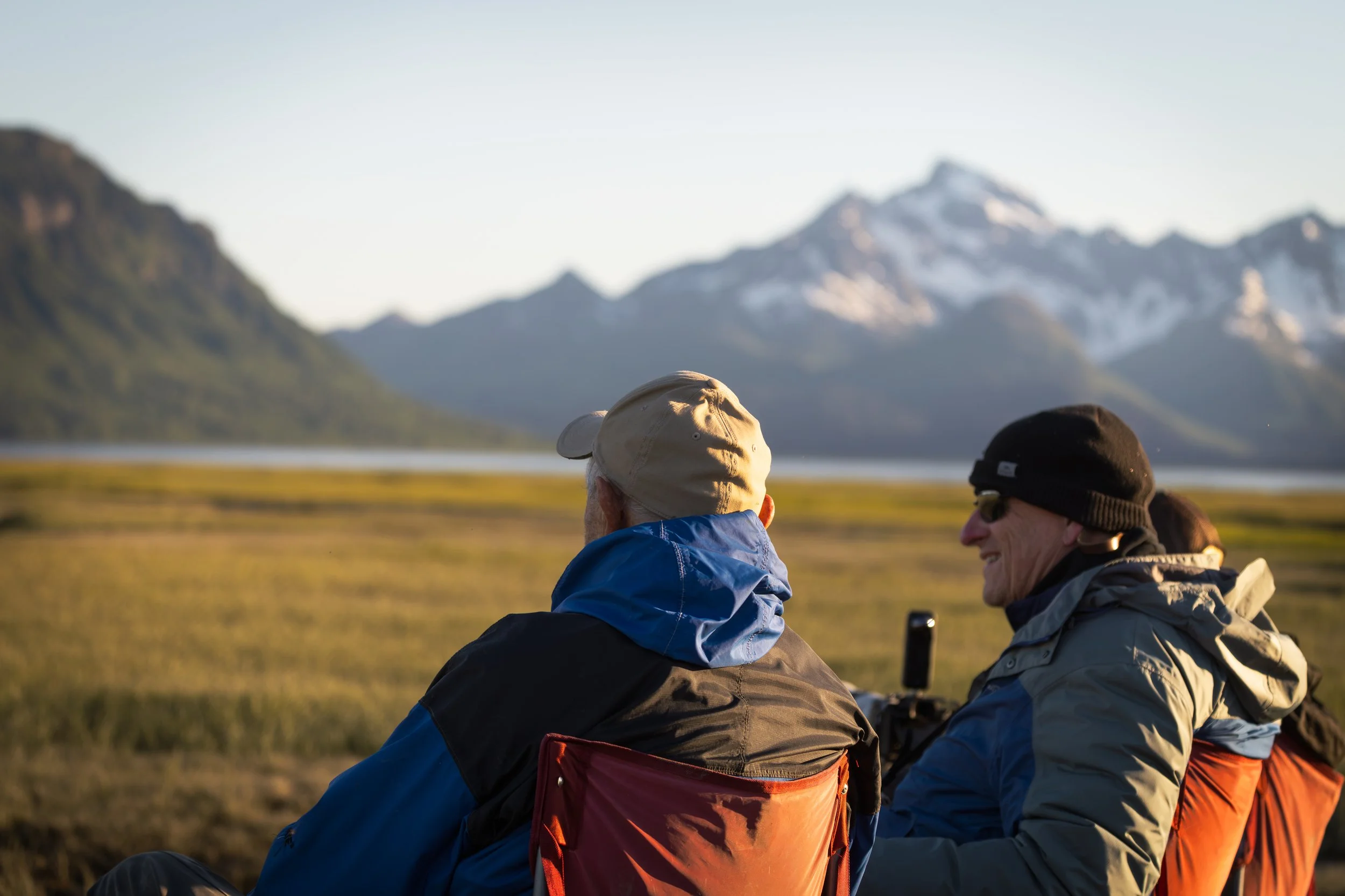 Two people sit outdoors with snow-capped mountains and a clear sky in the background, enjoying a scenic view.