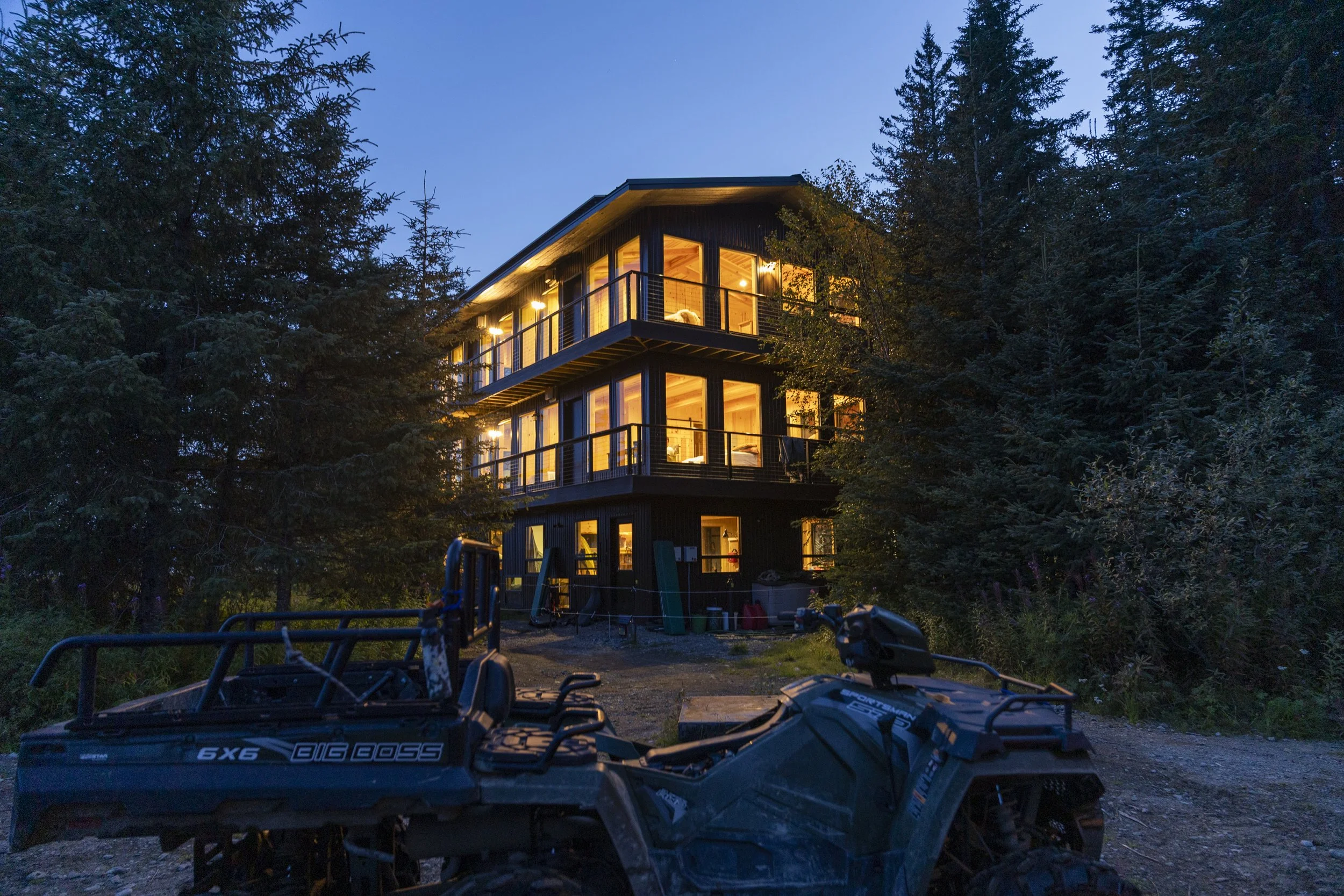 A modern multi-story house illuminated from within, surrounded by tall trees at dusk.