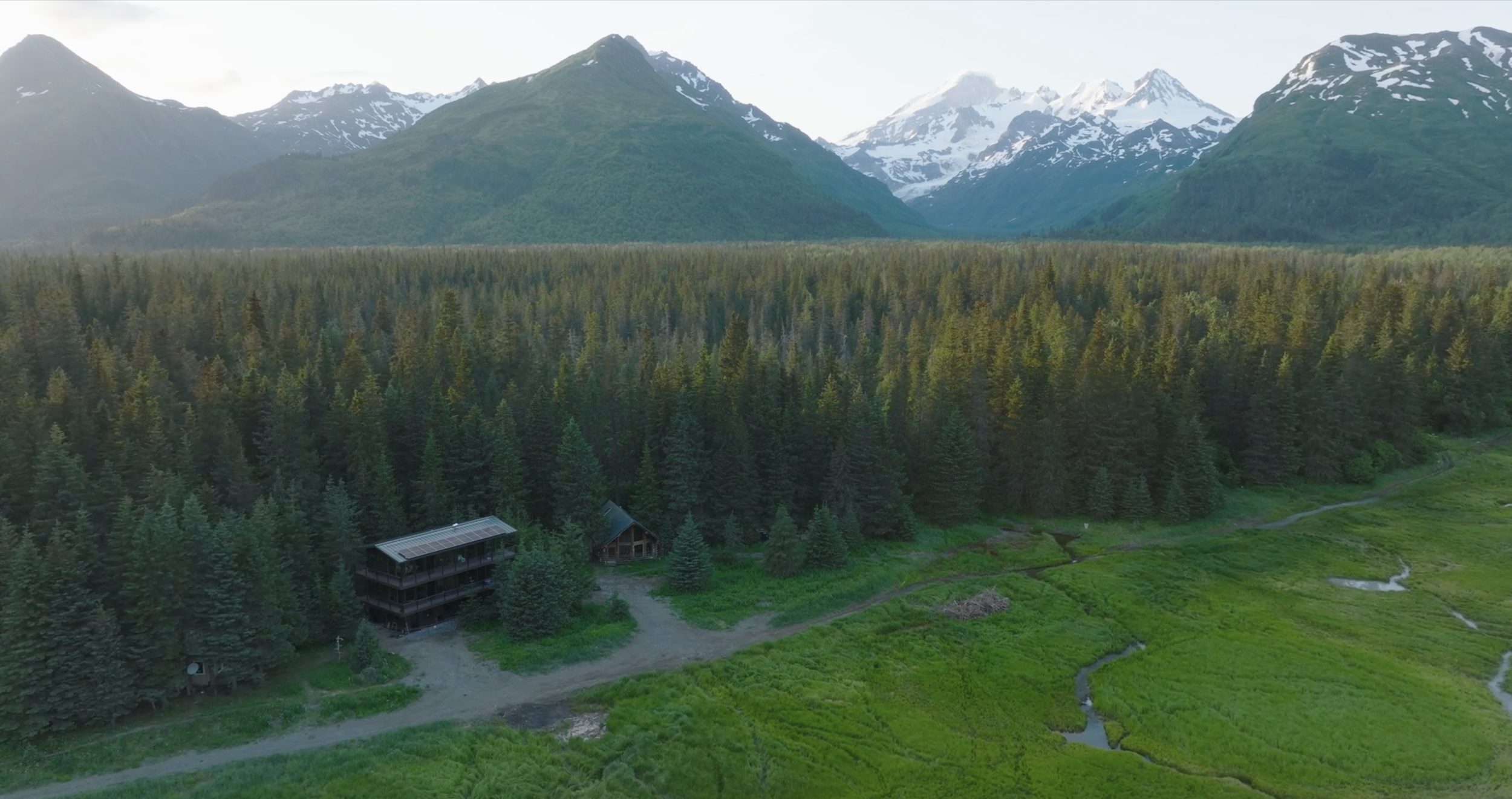 Aerial view of a dense forest with two small buildings and a winding path, surrounded by mountains with snow patches in the background.