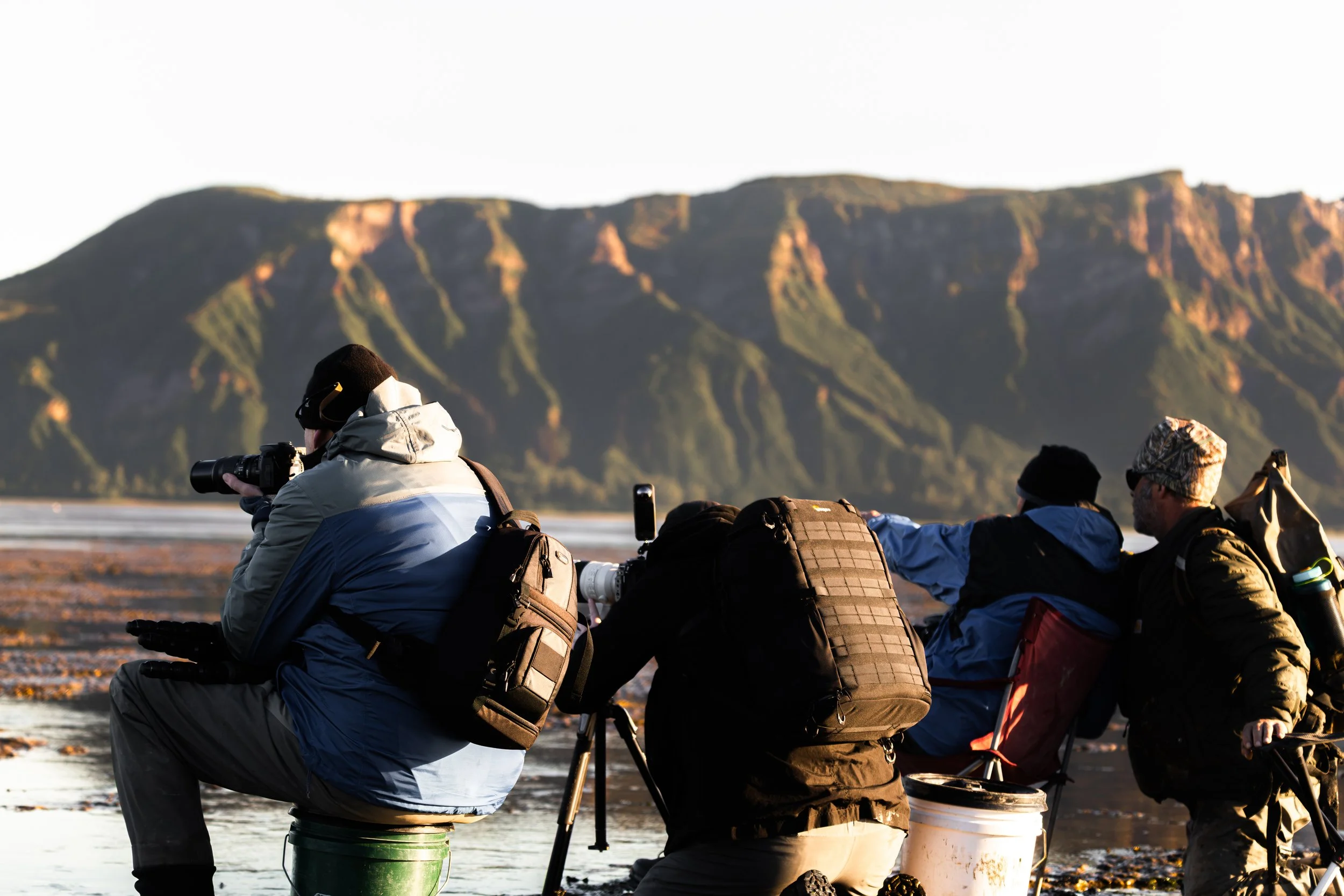 Group of people sitting outdoors with cameras, mountains in the background.