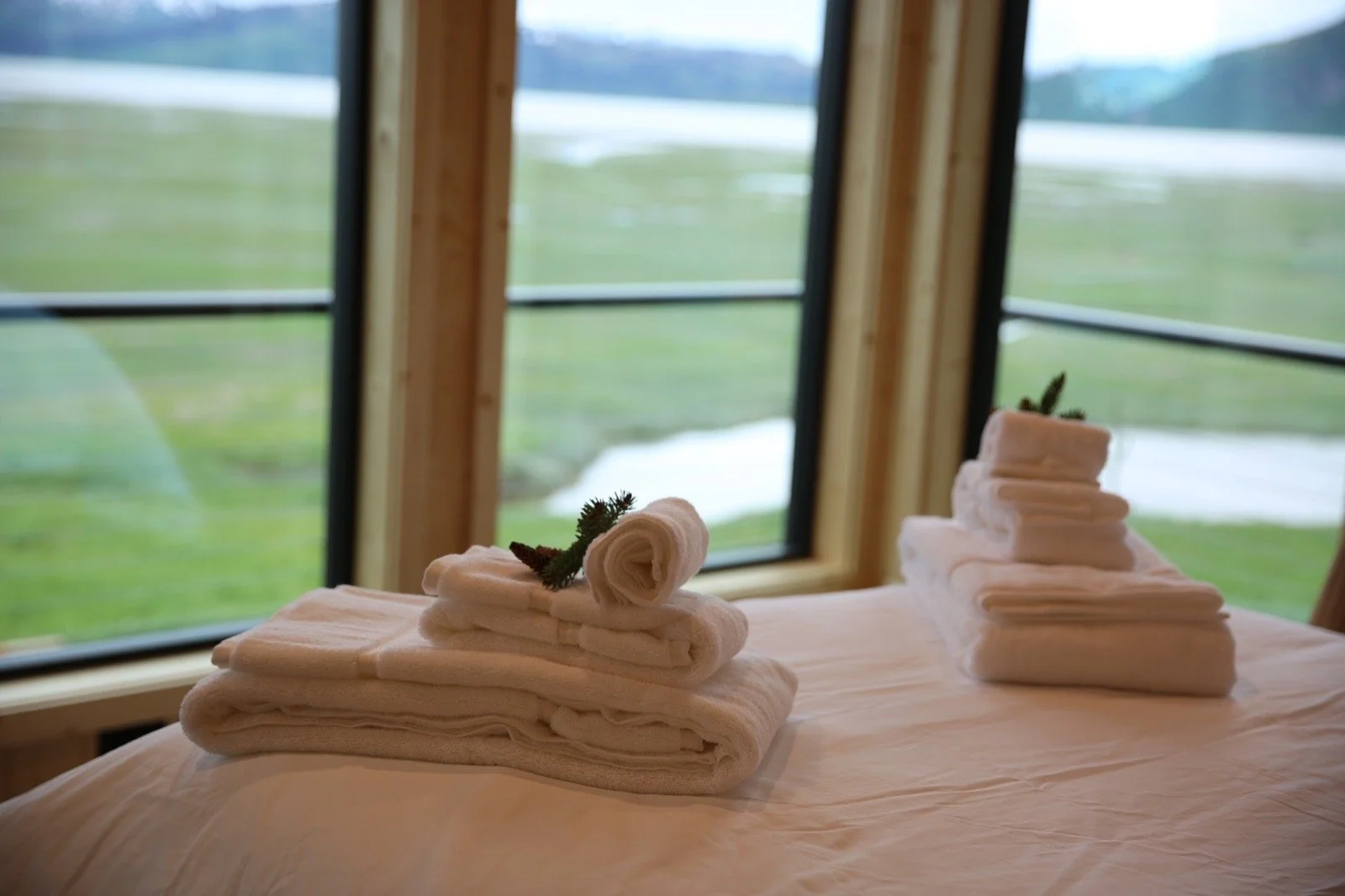 A bed with folded white towels arranged in a stack, decorated with a small sprig of greenery, placed in front of a large window overlooking a green landscape with patches of water.