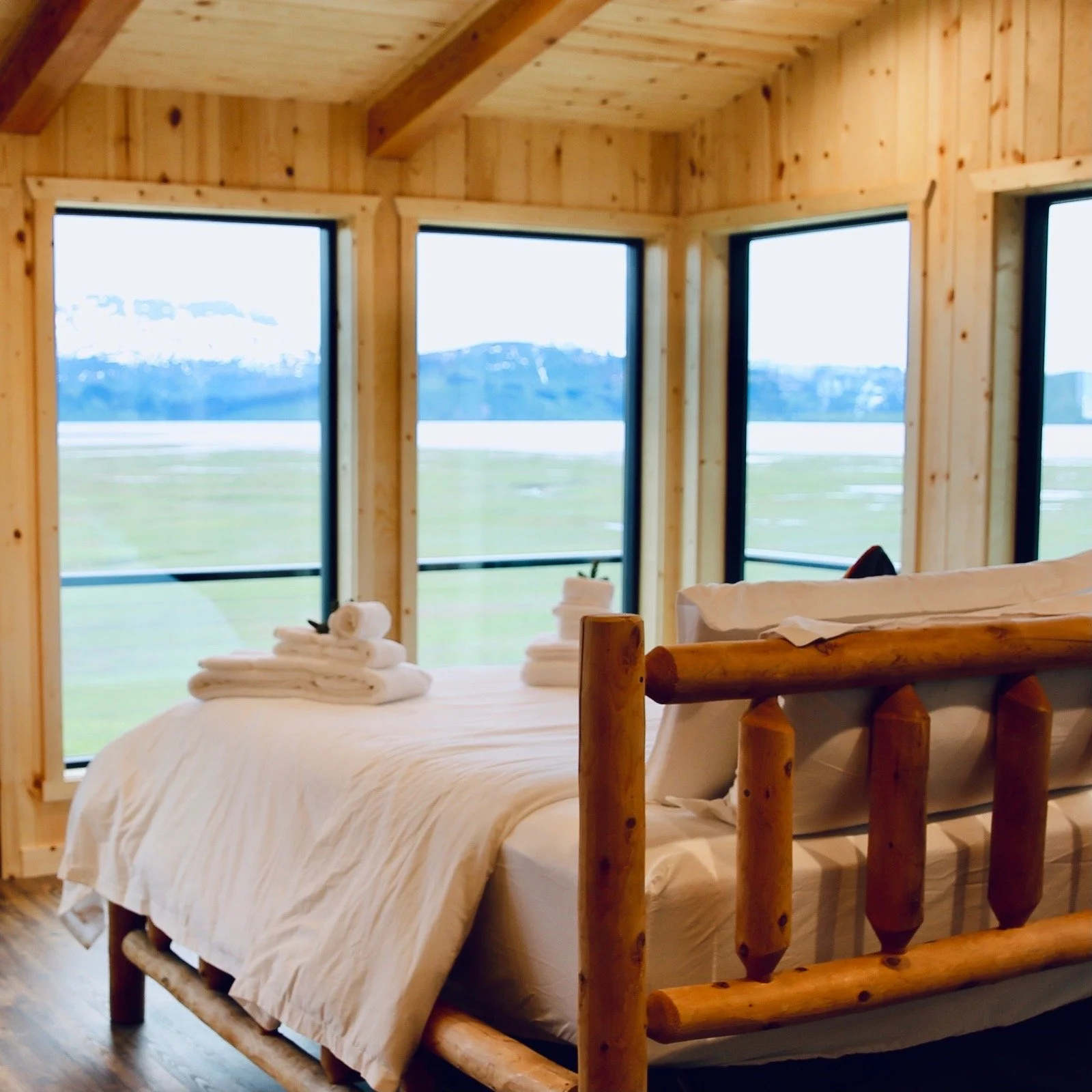 A cozy bedroom with wooden walls and ceiling, large windows offering a scenic view of distant mountains and fields, a bed with white bedding and neatly folded towels, and a rustic log headboard.
