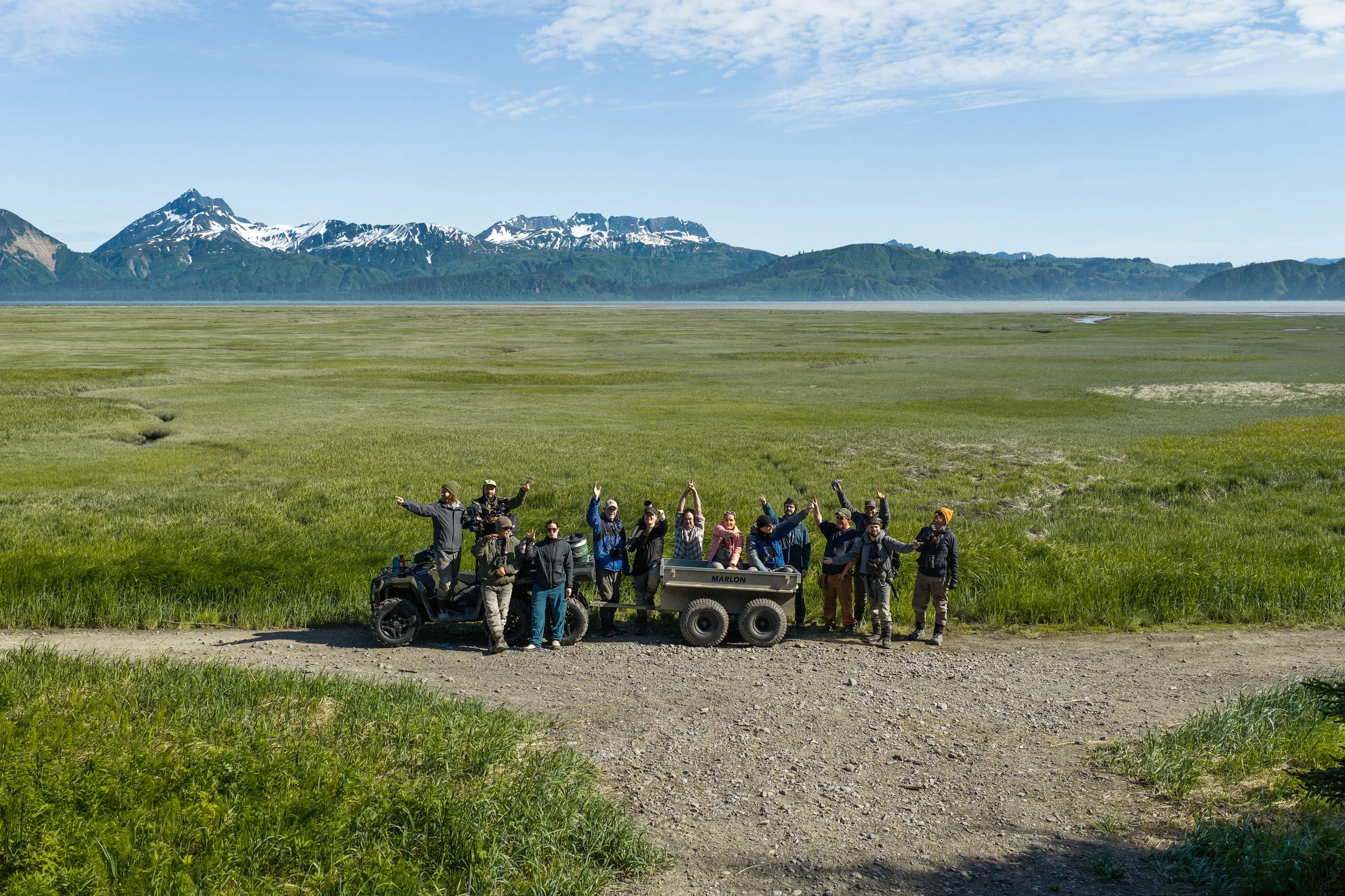 Group of people posing outdoors in a grassy field with mountains and a lake in the background, some waving and standing near a small motorcycle and trailer.
