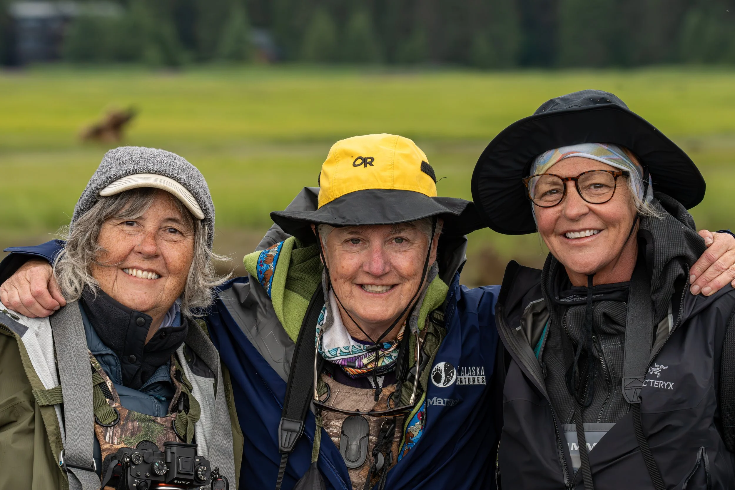 Three smiling women in outdoor gear, photo taken during daytime with a blurred green field and a dog in the background.