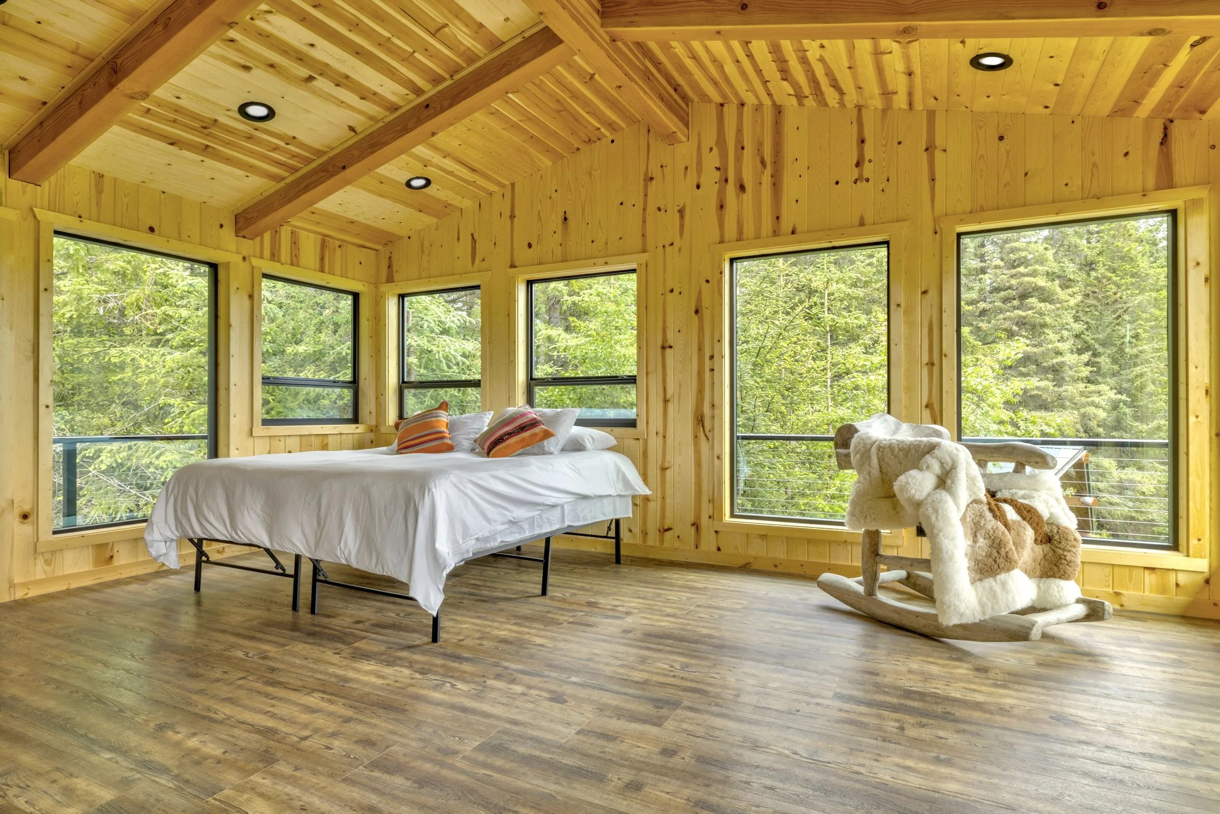 A cozy bedroom with wooden walls and ceiling, large windows showing lush greenery outside, a bed with white bedding and colorful pillows, and a rocking chair with plush throws.