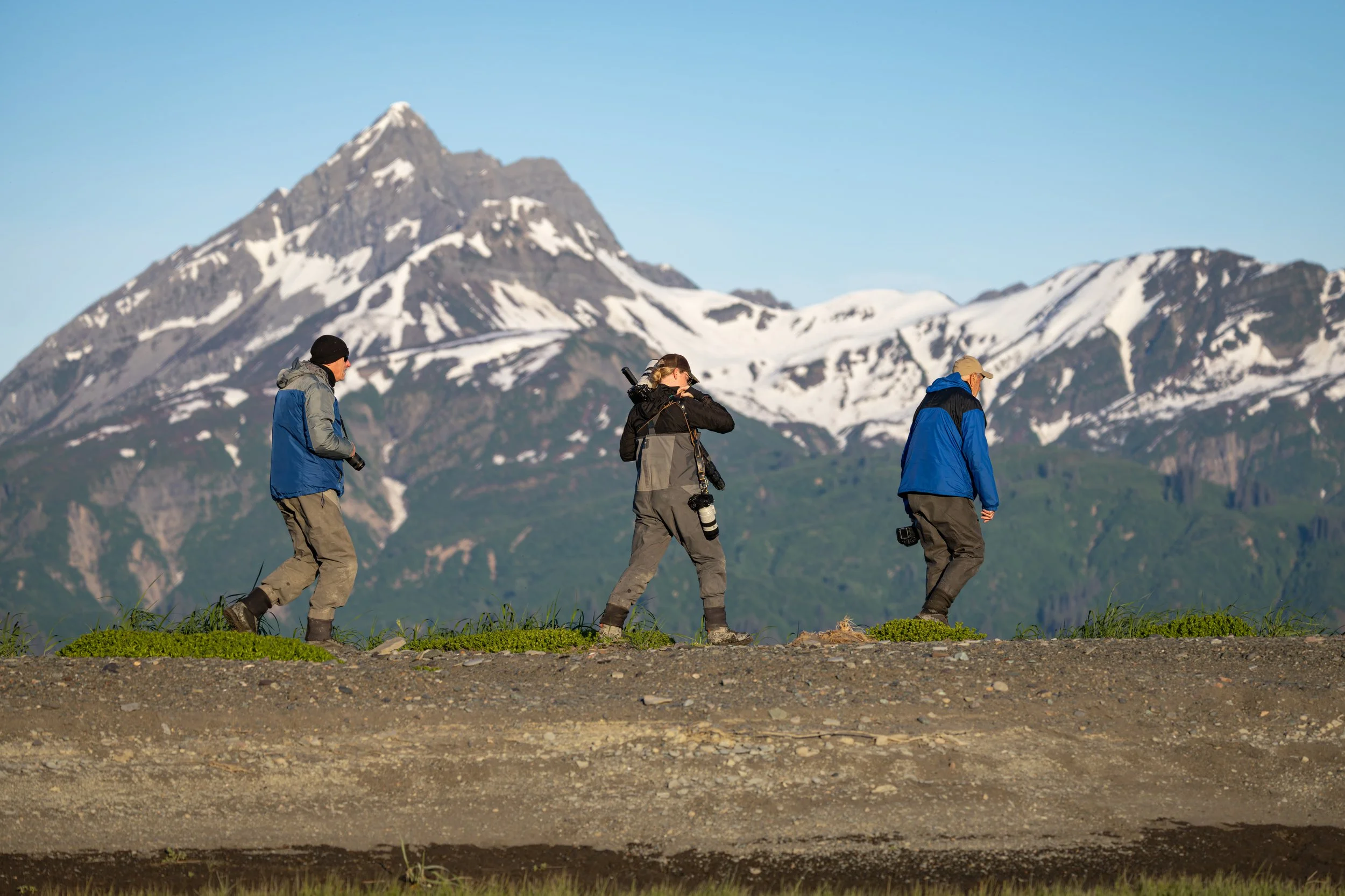 Three hikers walking across a grassy area with a snow-capped mountain in the background.