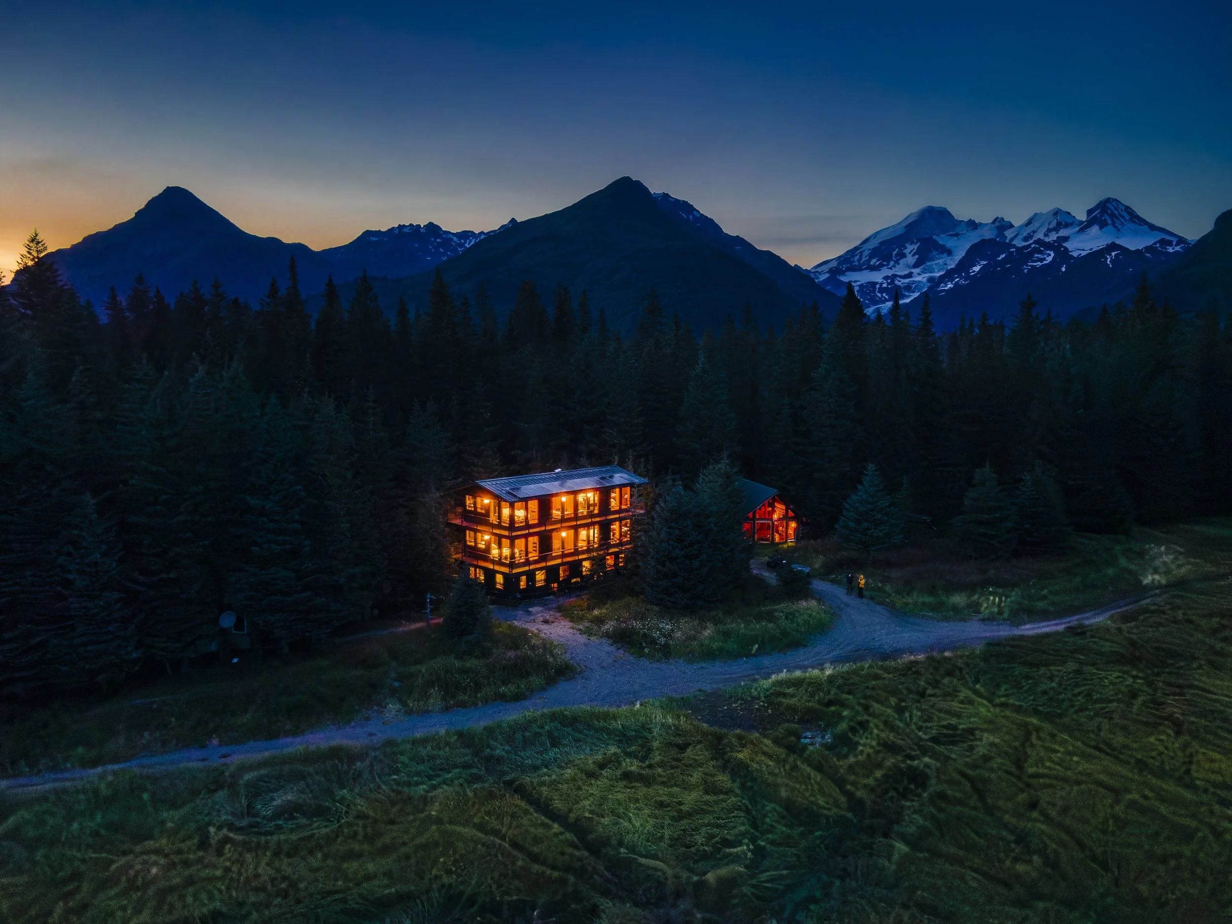 A mountain range at dusk with snow-capped peaks, a clear sky, and a forest of evergreen trees in the foreground. There are two houses with lit windows nestled in the trees.