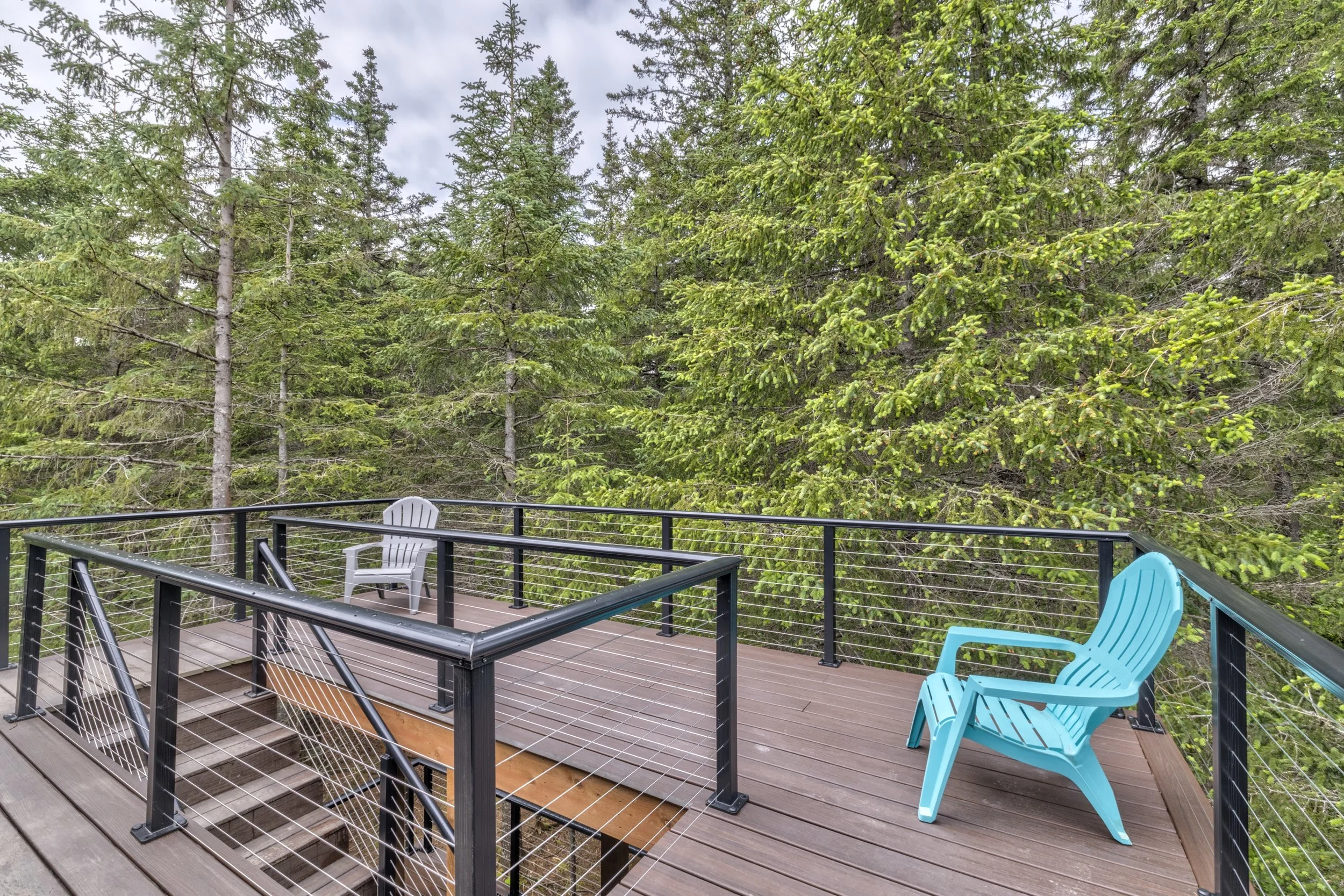 A wooden deck with a black metal railing and deck chairs, including a white plastic chair and a bright blue Adirondack chair, surrounded by green trees with dense foliage.