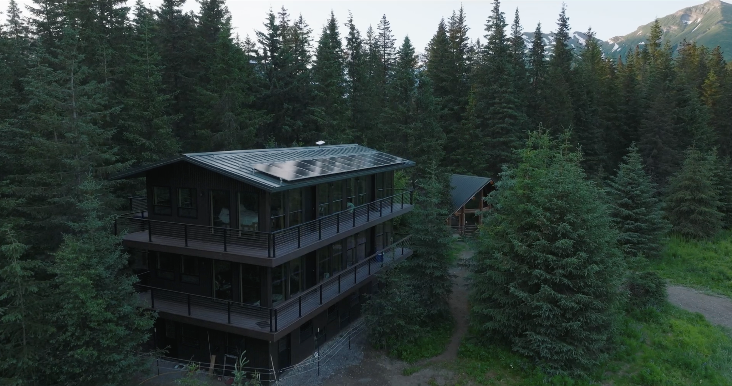 A modern house with solar panels on the roof, surrounded by tall pine trees in a mountainous forest area.