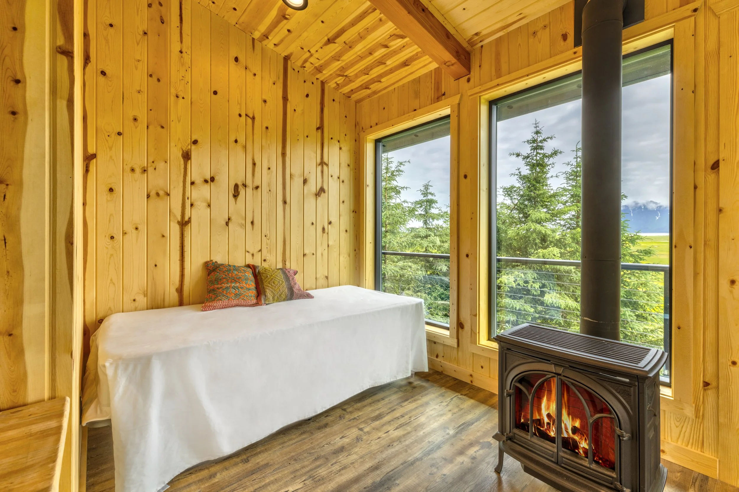 Interior of a cozy, rustic wooden cabin with large windows showing trees outside, a white bed with colorful pillows, and a black fireplace with a lit fire.