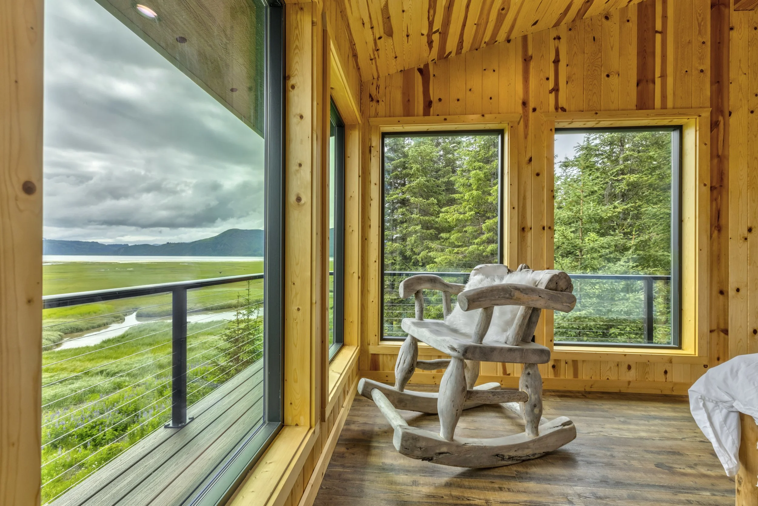 Wooden interior room with large windows showing greenery and mountains outside, rustic wooden chair with white sheepskin cushion.