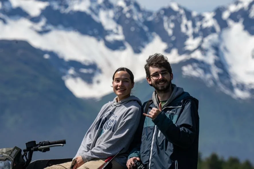 Two people smiling outdoors with snow-capped mountains in the background, one on a motorcycle.