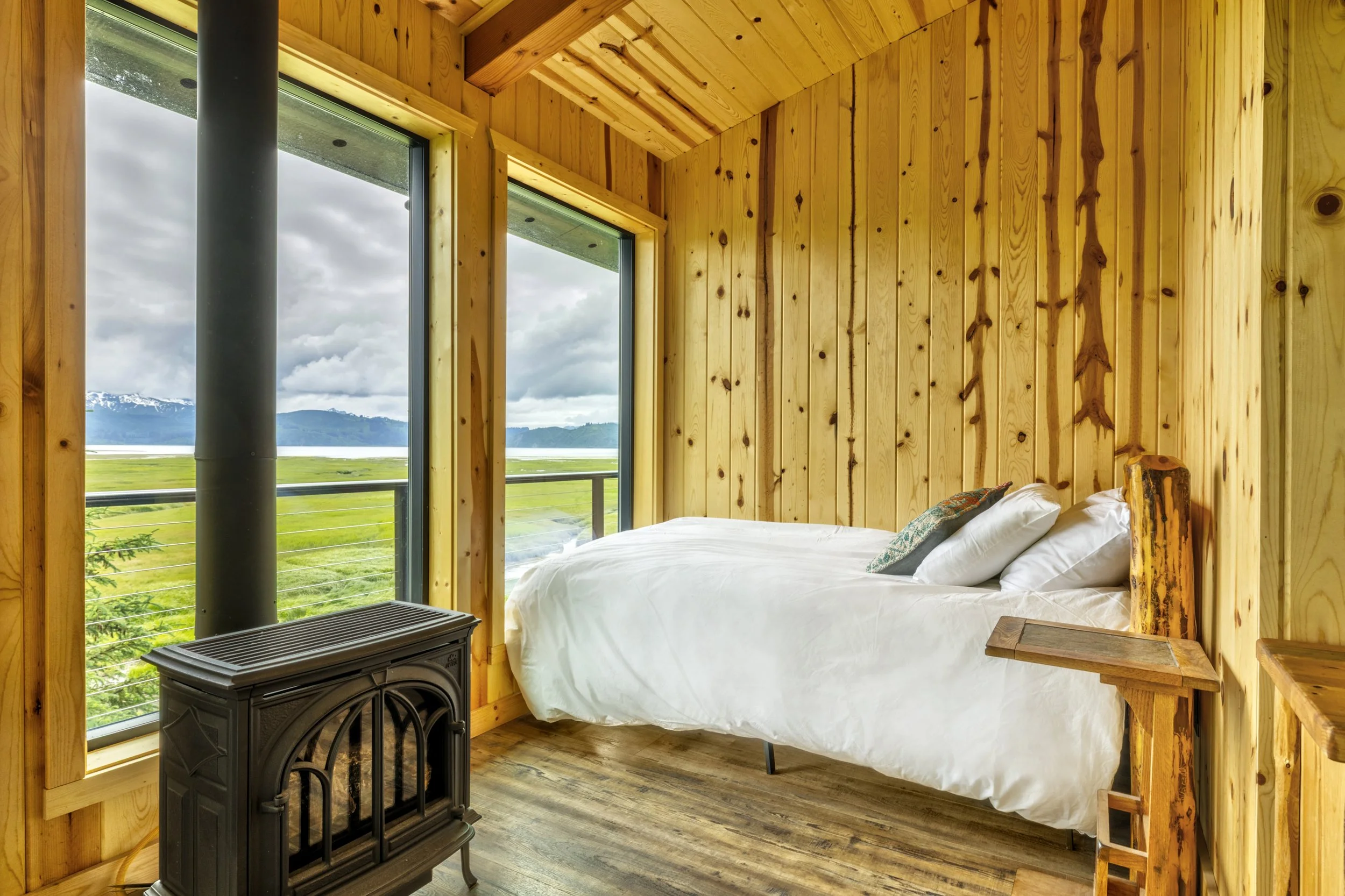 A cozy bedroom with wood-paneled walls and ceiling, a large window showing a scenic landscape with mountains, water, and cloudy sky, a bed with white sheets, a small wooden side table, and a black cast iron stove.