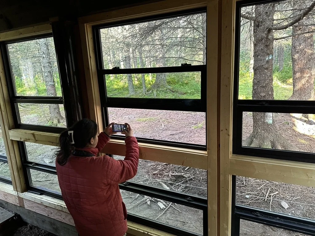 A woman in a red jacket taking a photo of a forest scene through a multi-pane window in a wooden structure.