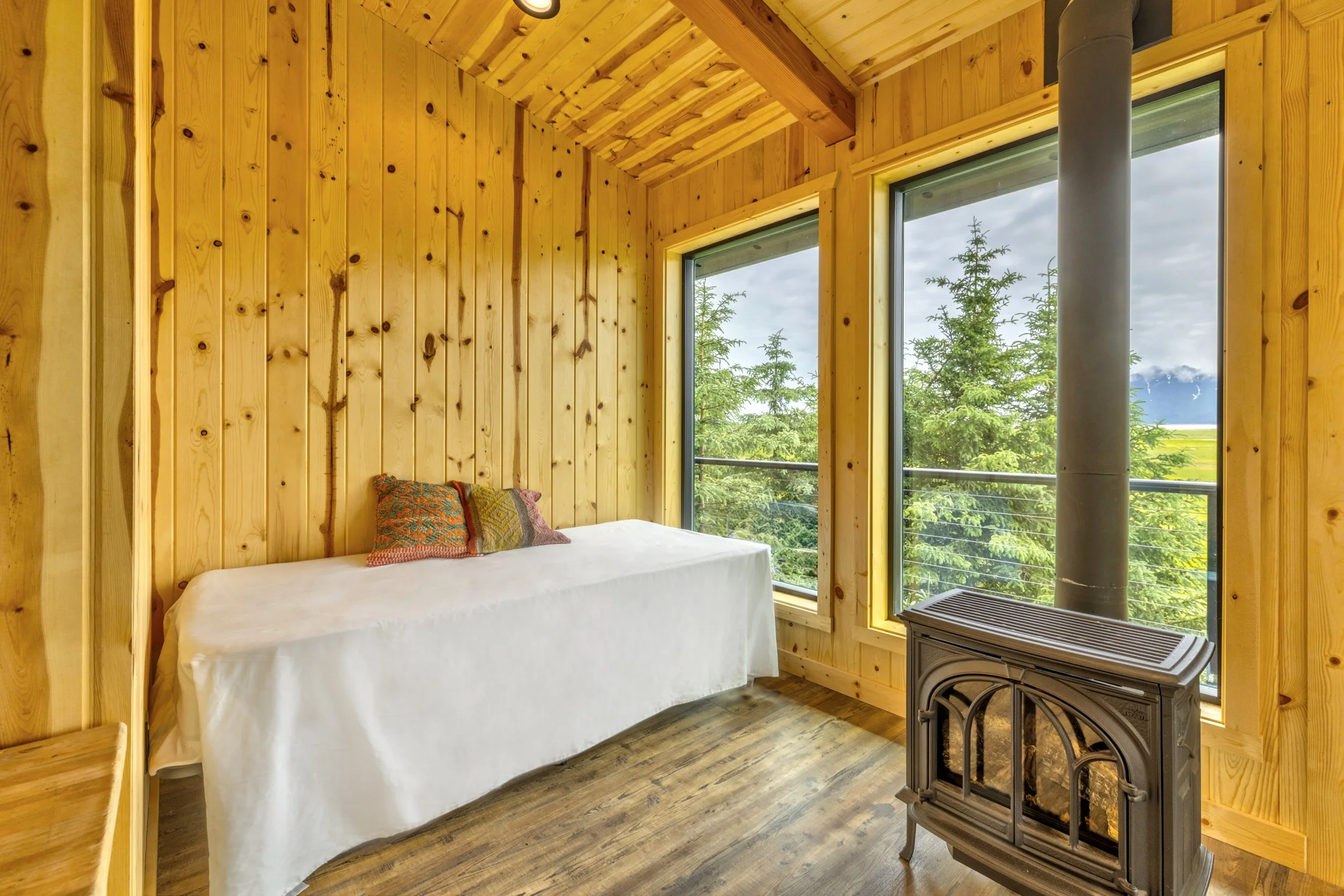 The image shows a cozy room with wood-paneled walls and ceiling, large windows overlooking trees and mountains, a bed with a white cover and colorful pillows, and a wood stove in the corner.