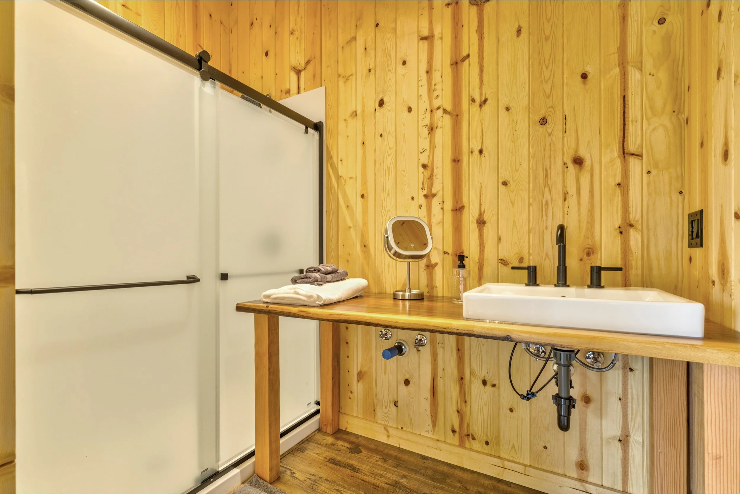 A bathroom with wooden wall paneling, a white sink with black fixtures, a small mirror, a soap dispenser, and folded towels on a wooden countertop.
