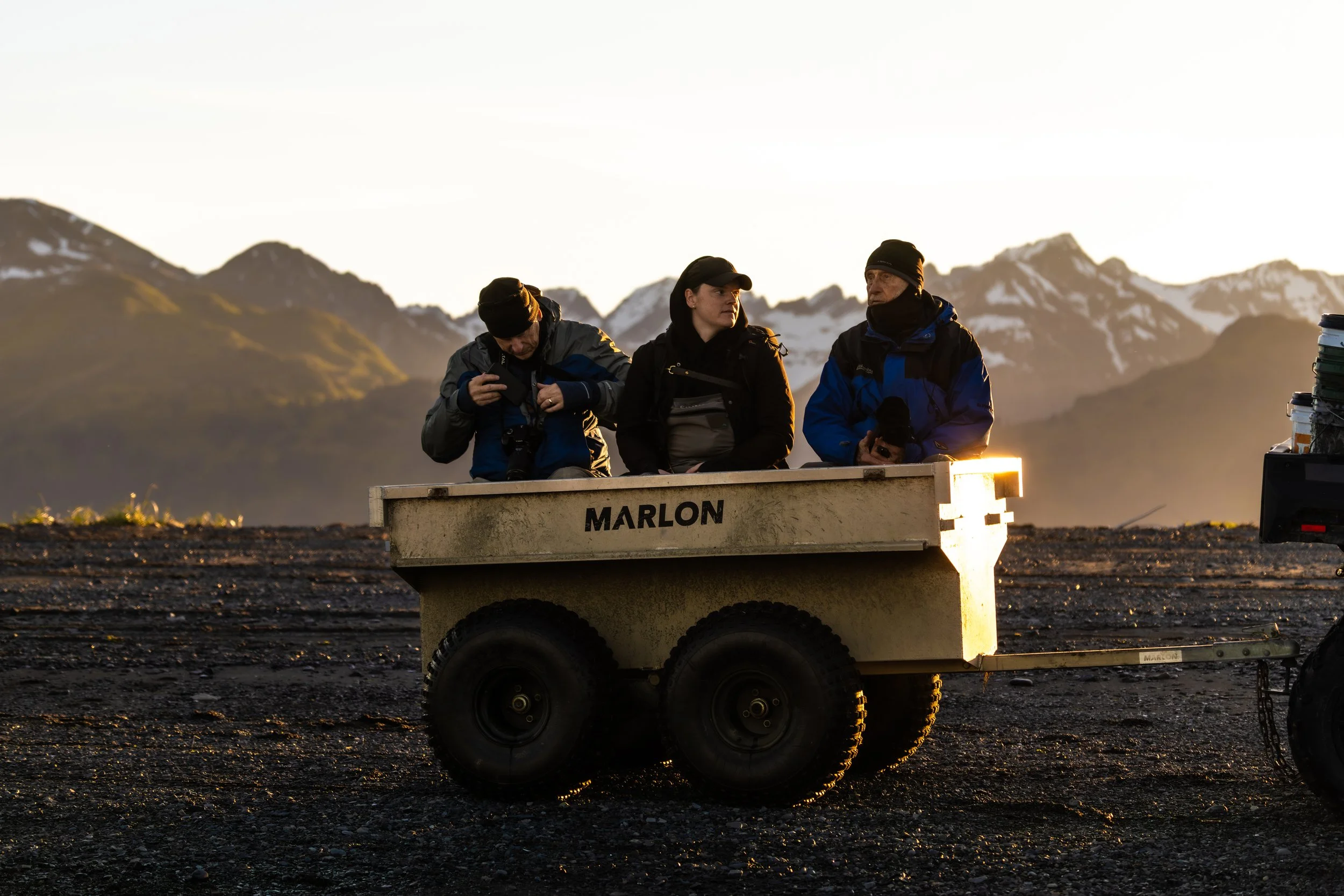 Three people sit on a trailer labeled 'Marlon' in a rugged outdoor setting with mountains in the background, possibly during sunrise or sunset, dressed warmly for cold weather.
