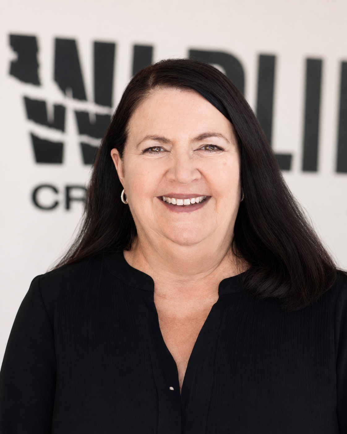 A smiling woman with dark brown hair wearing a black top, standing in front of a blurred logo that says 'Wildlings'