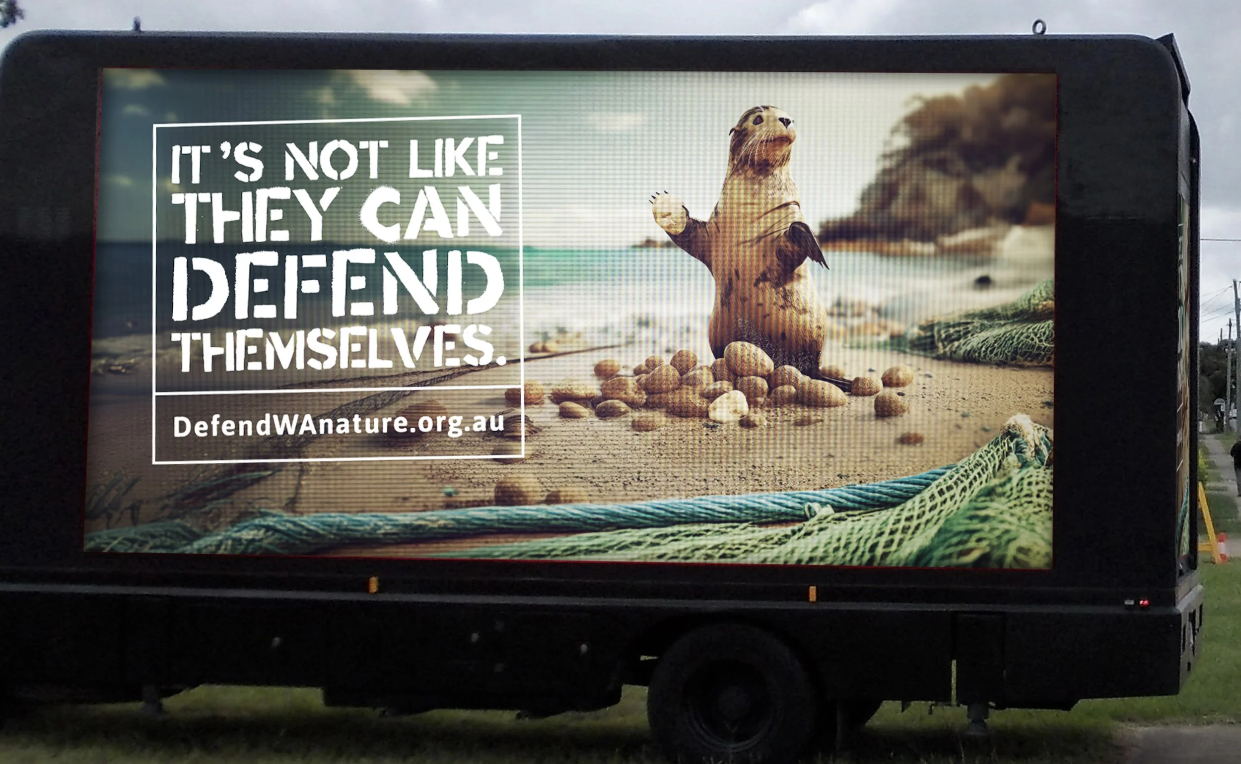 A large digital billboard displays an image of a beach scene with an animated otter holding a shell in its paw, surrounded by pebbles and a net in the foreground. The billboard features text that reads, 'It's not like they can defend themselves,' and