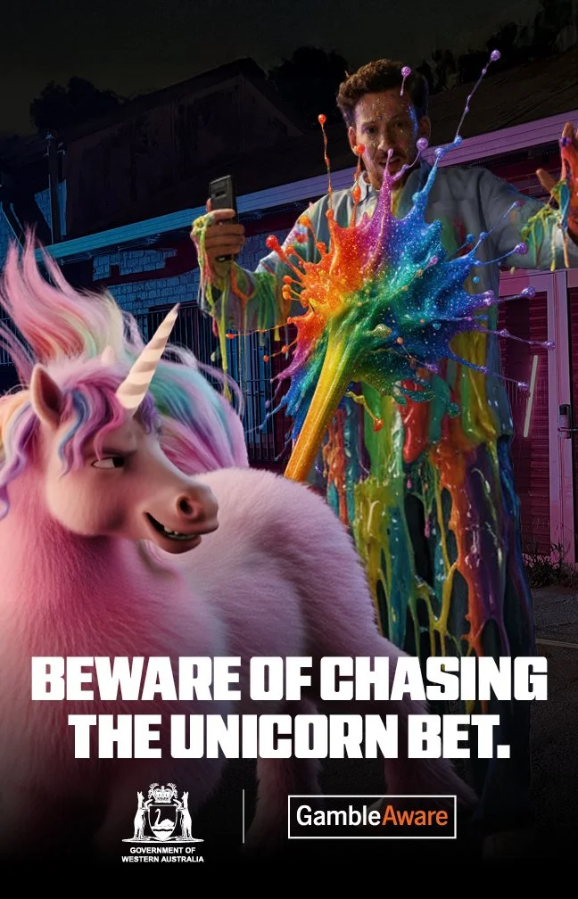 A person holding a phone in front of a colorful splash of paint and a cartoon unicorn. The image has a warning message about gambling, with logos from the Government of Western Australia and GambleAware.