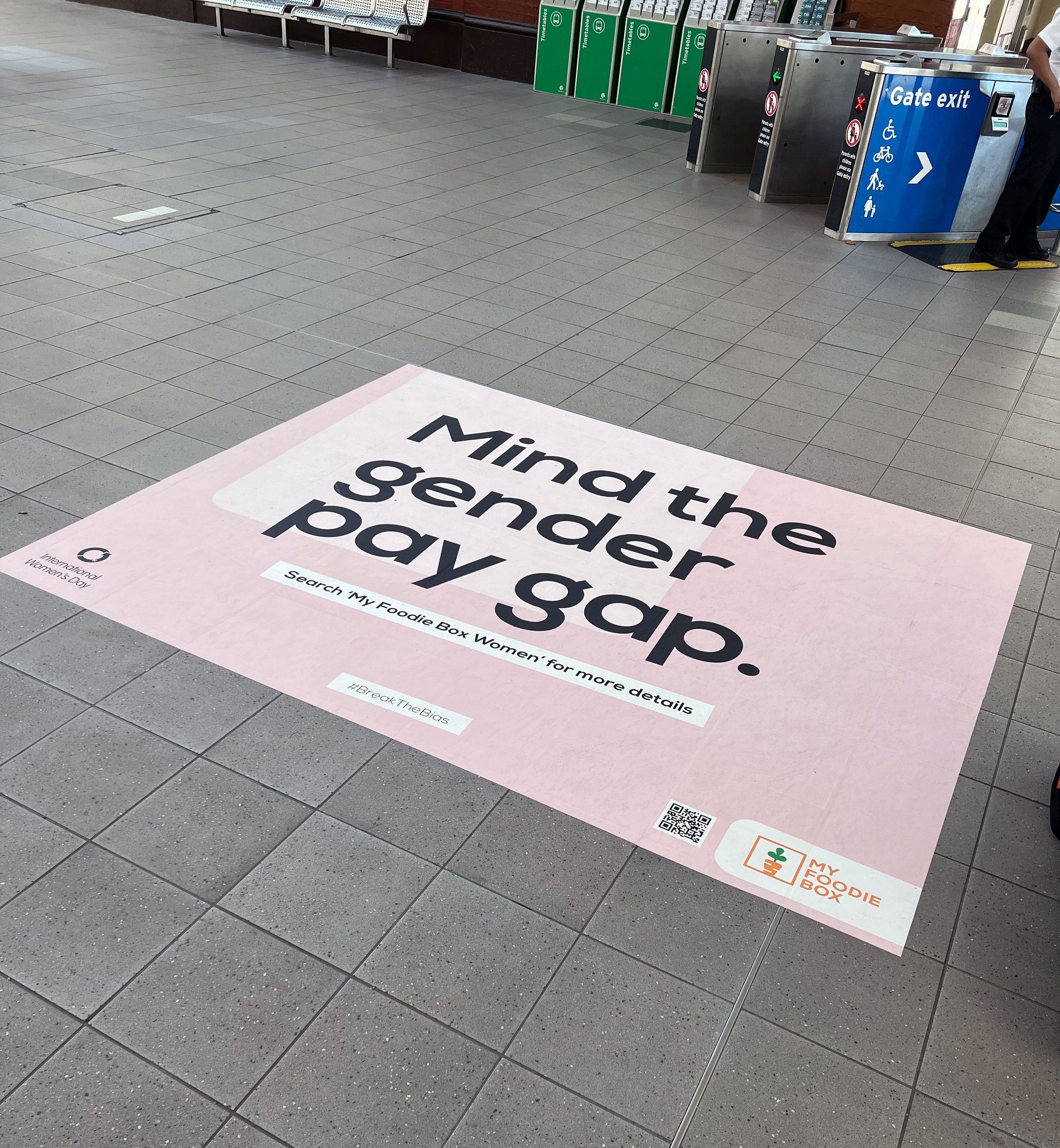 An airport or train station interior with a large pink social distancing floor sticker that reads 'Mind the sender policy' and a QR code. There are green timetable signs, metal turnstiles, and a blue gate exit sign with accessibility symbols includin
