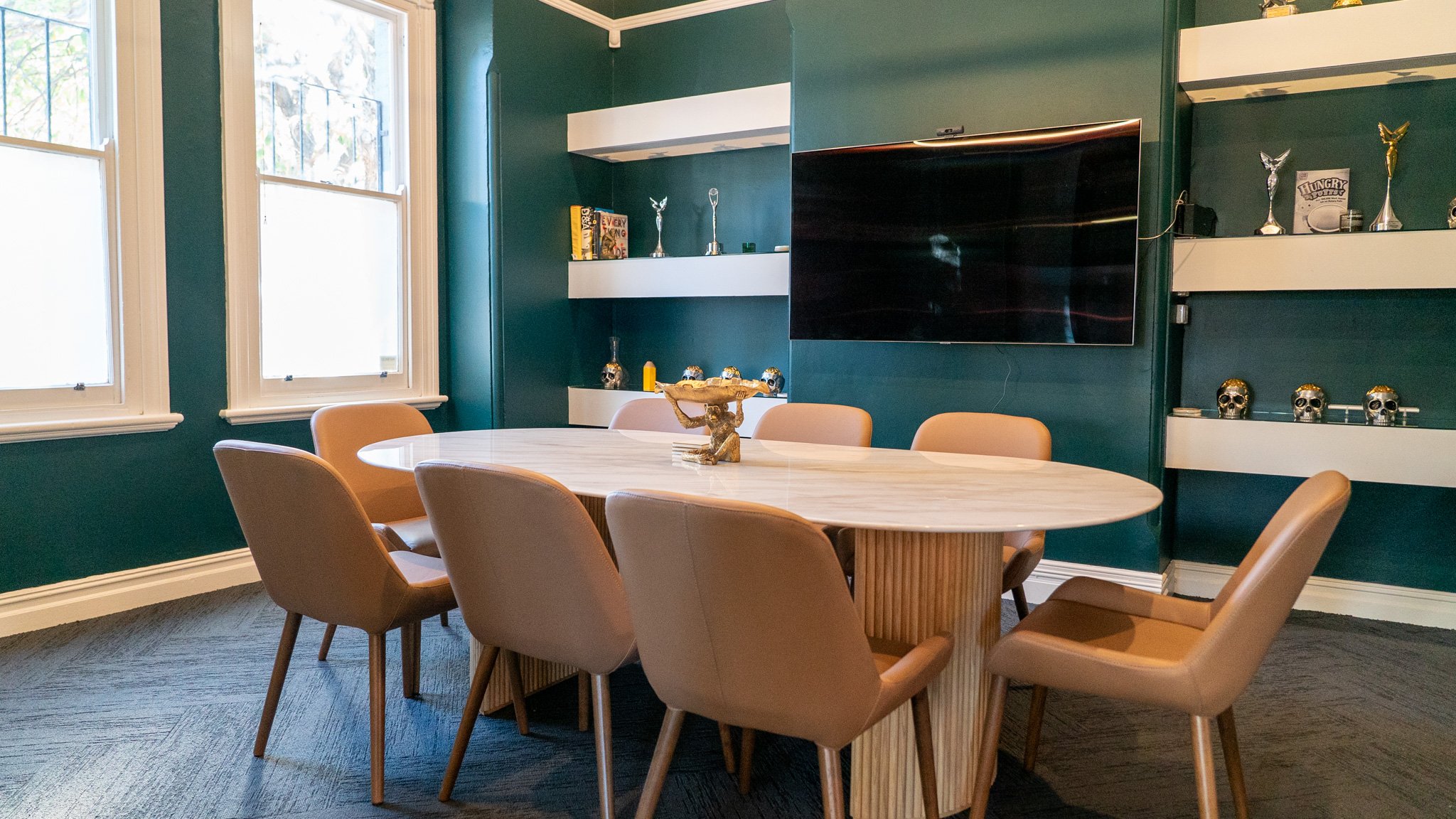 A dining room with a large, oval, wood table surrounded by beige chairs. There are three large windows on the left side, with white frames. The turquoise-colored walls have white floating shelves displaying various trophies and decorative items, including skulls with gold accents and small statues. A flat-screen TV is mounted on the wall in the background.