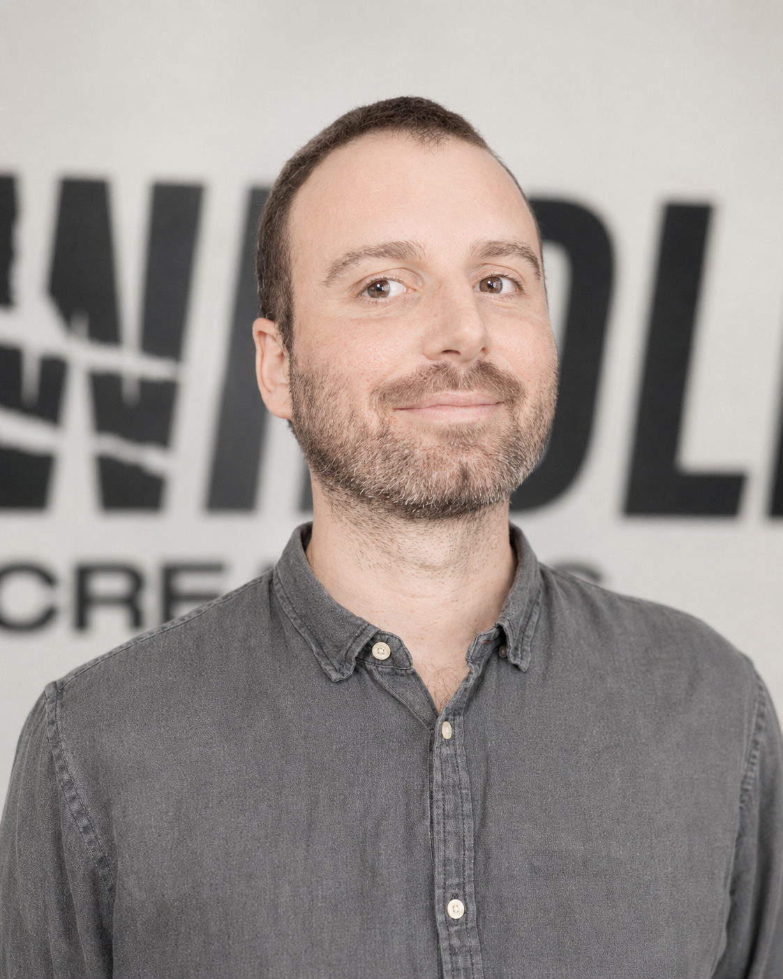 A man with short brown hair, a beard, and a mustache, wearing a gray button-up shirt, standing in front of a wall with partially visible black and white text.