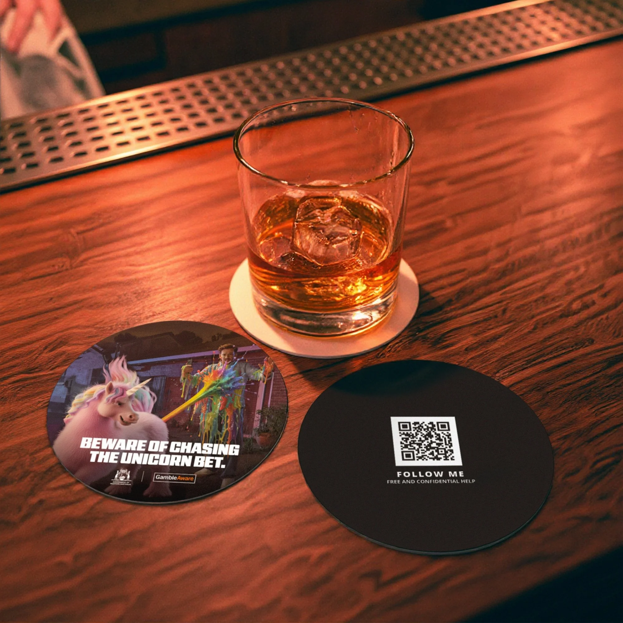 A close-up of a glass of whiskey on a wooden bar counter, with a coaster featuring a unicorn and a warning about chasing the unicorn bet, and a QR code on a black square next to the glass.