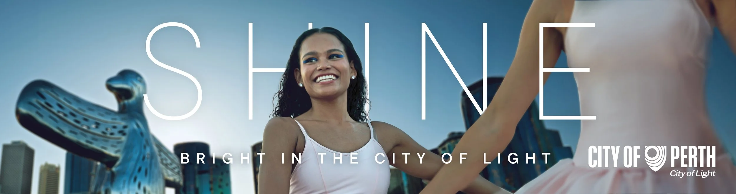 Smiling woman with dark curly hair and bright makeup standing outdoors in front of city buildings and city of Perth logo, with a large metallic sculpture of a bird in the background.
